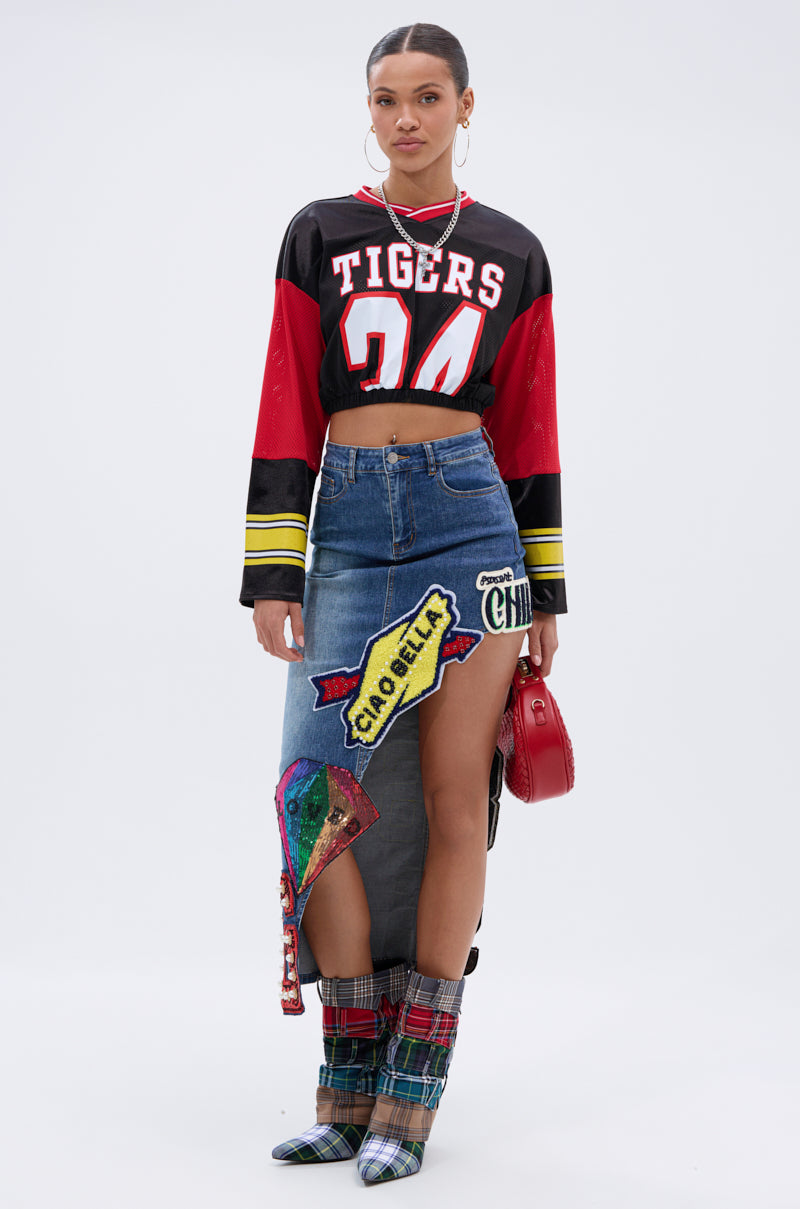 A woman poses in a cropped “Tigers 24” jersey, plaid boots, hoop earrings, a small red purse, and the VARSITY SQUAD DENIM GRAPHIC MIDI SKIRT with bold patterns and a high slit against a plain background. 7