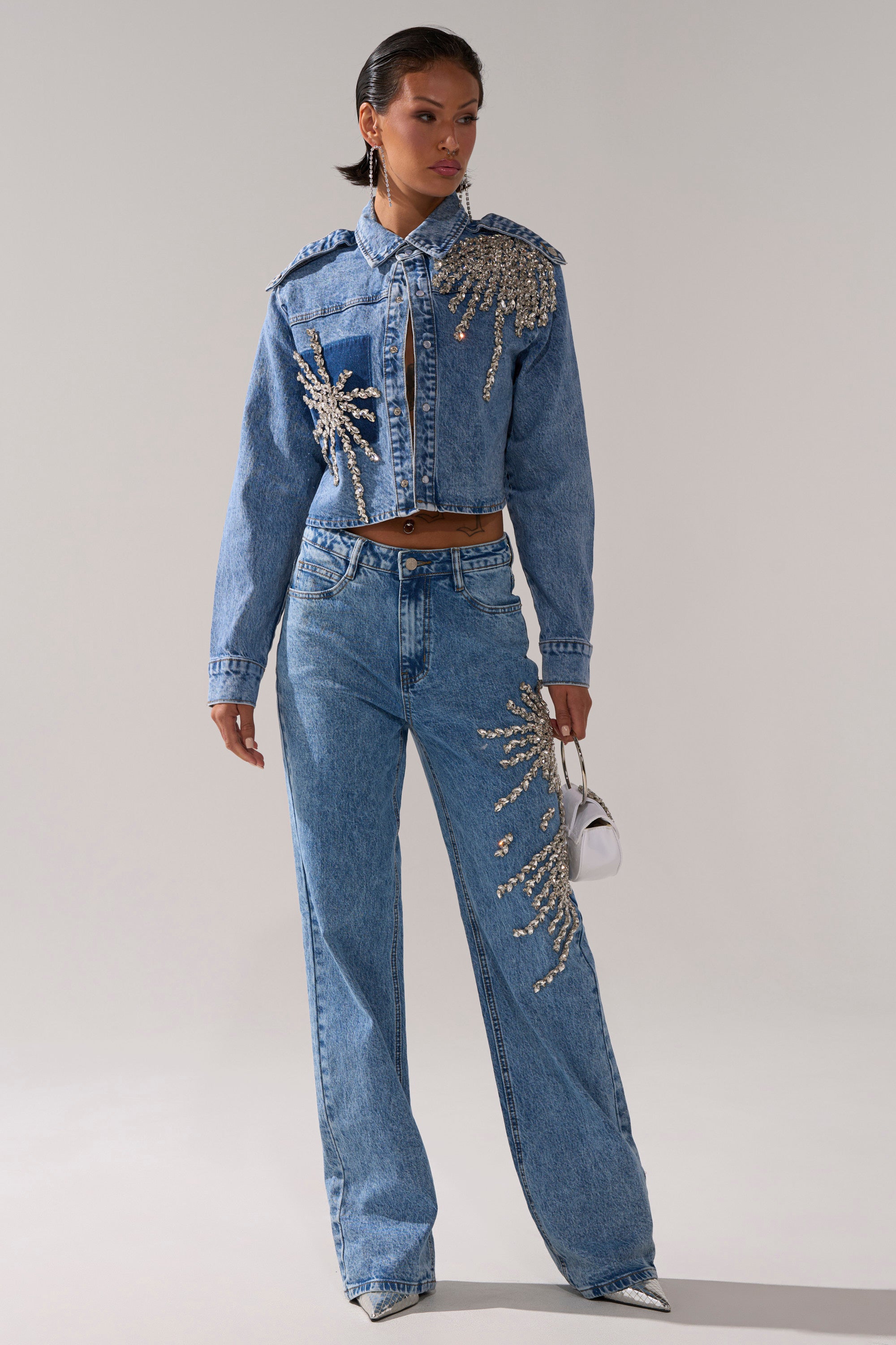 Wearing the SPARKLE AND SHINE QUEEN RELAXED DENIM jacket and high-rise jeans, a person holds a small white handbag while standing against a plain backdrop.