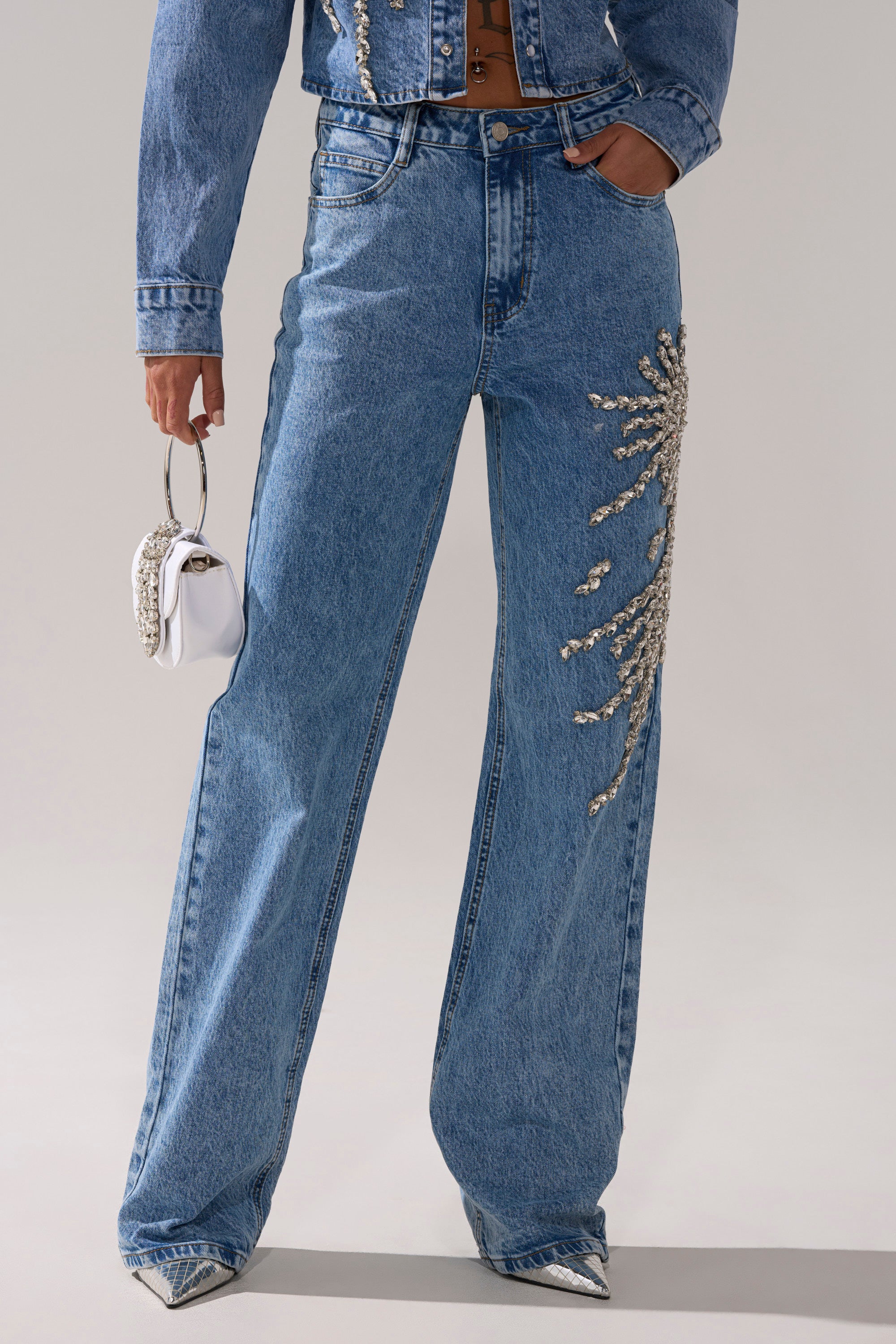 A person wears the SPARKLE AND SHINE QUEEN RELAXED DENIM set—high-rise, wide-leg jeans and a matching jacket with rhinestone sunburst embellishments—paired with silver pointed-toe shoes and a small white handbag.