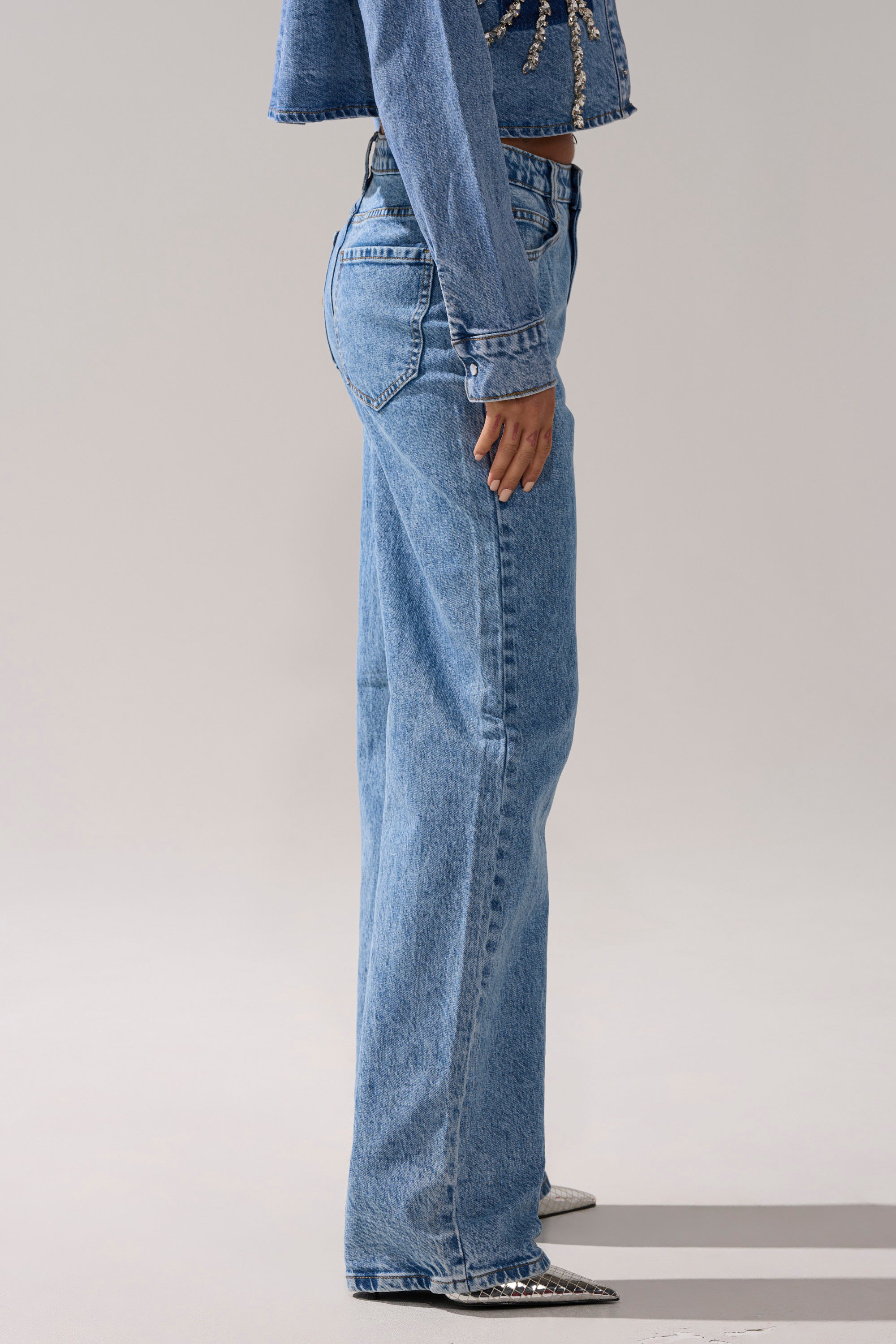 A person wearing SPARKLE AND SHINE QUEEN RELAXED DENIM jeans and a matching jacket stands in profile against a plain light background, with only part of the jacket and the lower legs and feet visible.