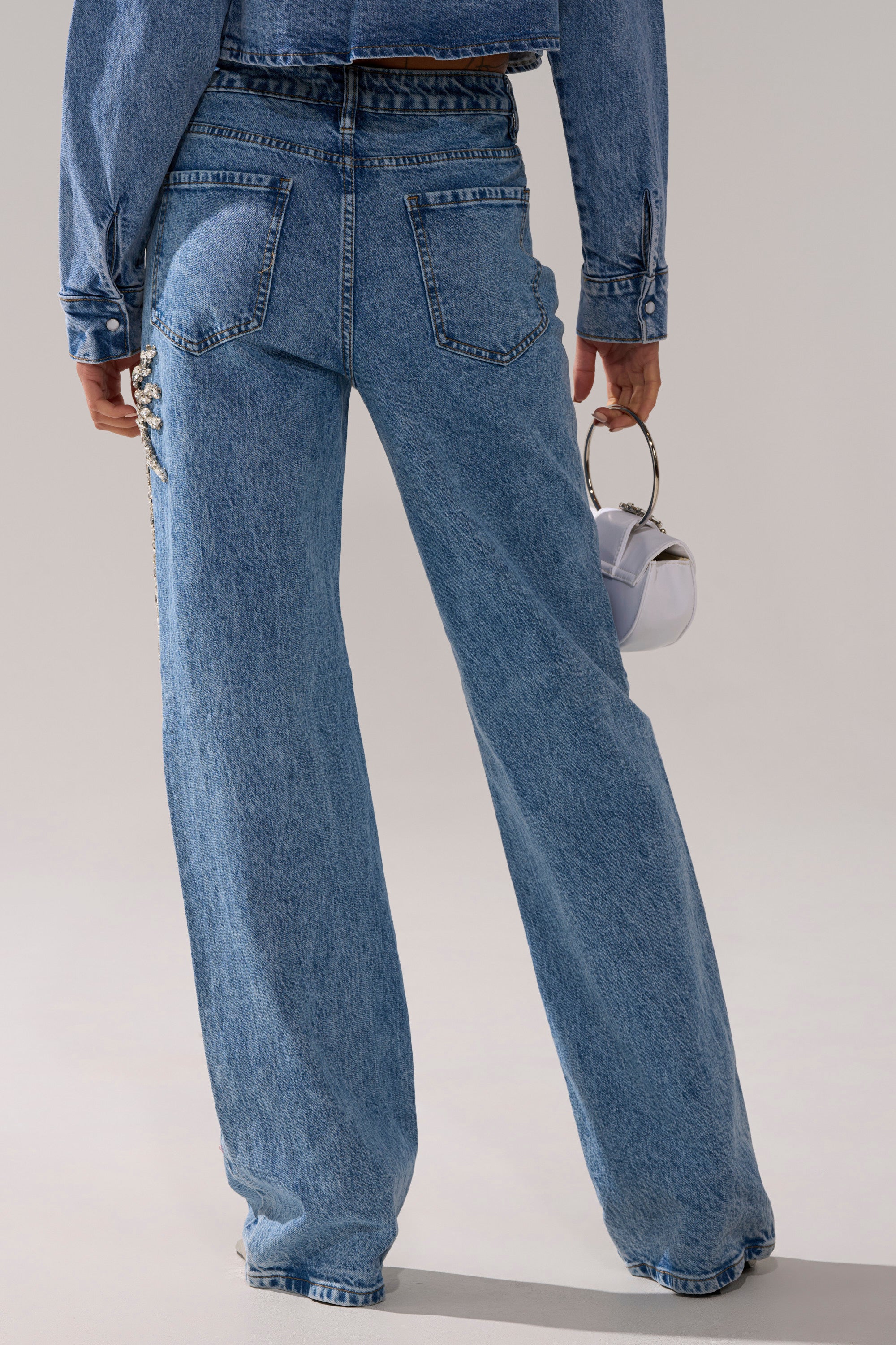 A person wearing SPARKLE AND SHINE QUEEN RELAXED DENIM jeans and a matching jacket holds a small white handbag. The photo, taken from behind, captures only the lower body.