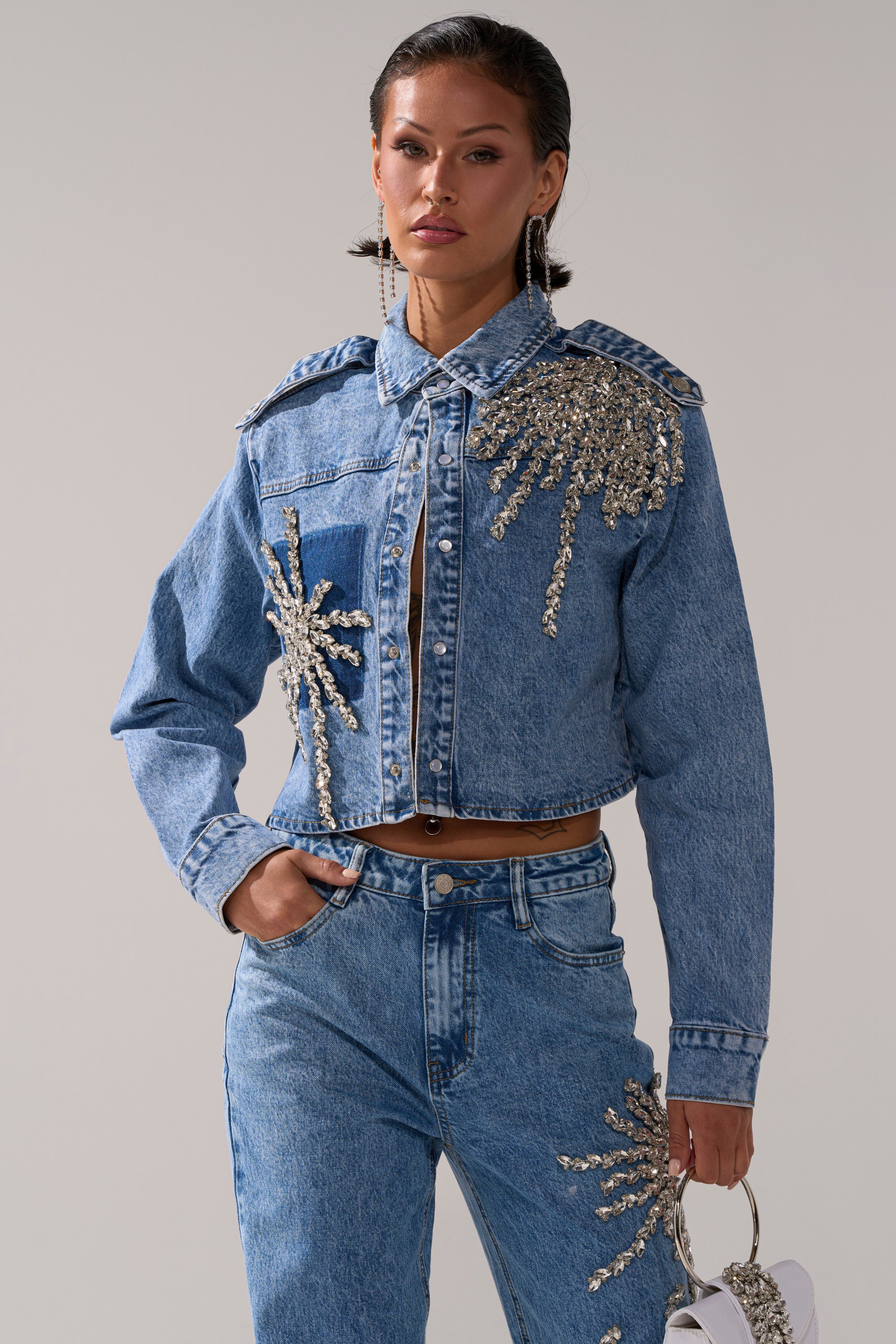 A woman wears the SPARKLE AND SHINE QUEEN DENIM BUTTON DOWN, shimmering with rhinestone embellishments, paired with jeans and a small bag, one hand in her pocket, against a plain background.