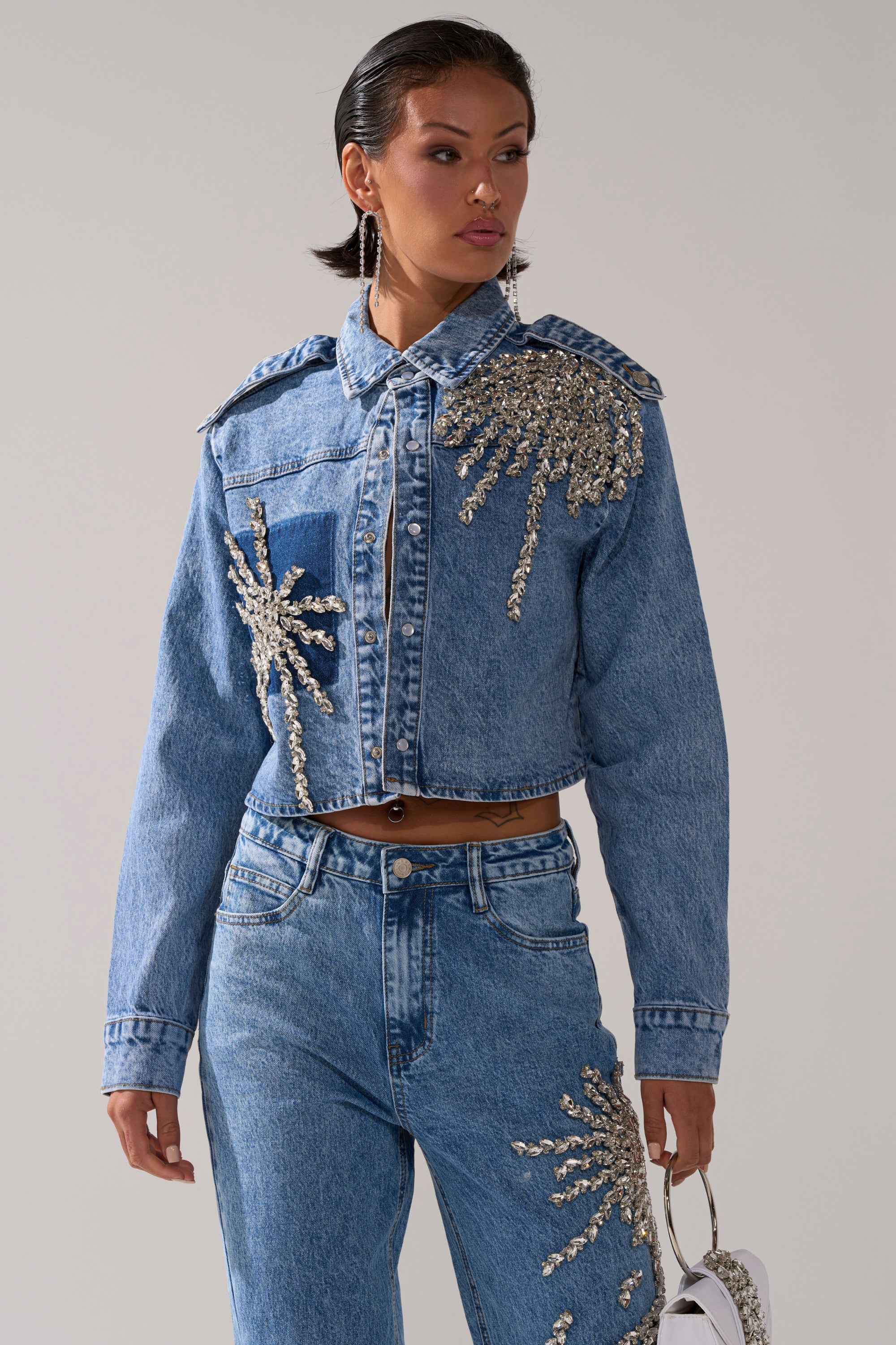 A woman in the SPARKLE AND SHINE QUEEN DENIM BUTTON DOWN and silver-beaded jeans holds a white handbag, standing against a plain light background.