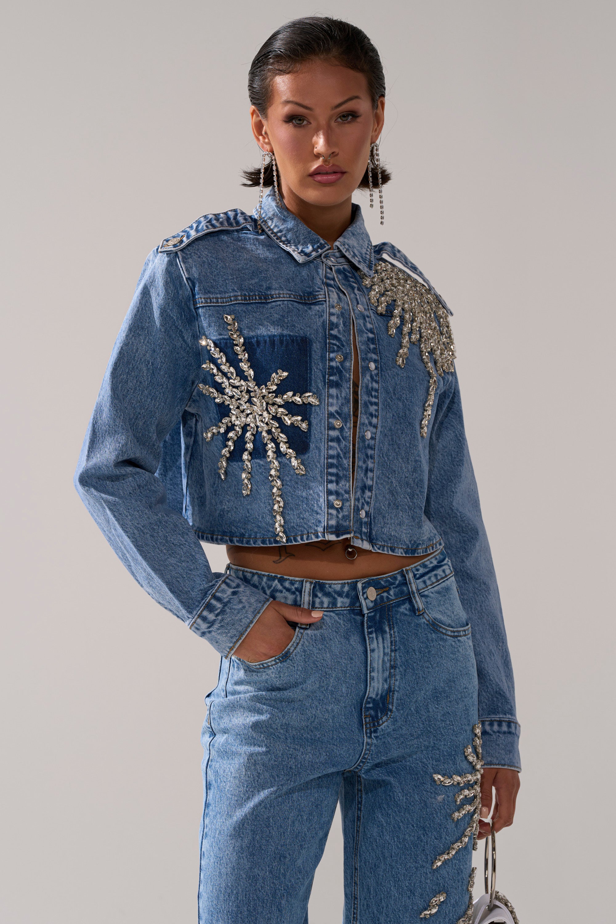 Wearing the SPARKLE AND SHINE QUEEN DENIM BUTTON DOWN, adorned with large sparkling silver rhinestones, a person stands against a plain background with one hand in their pocket.