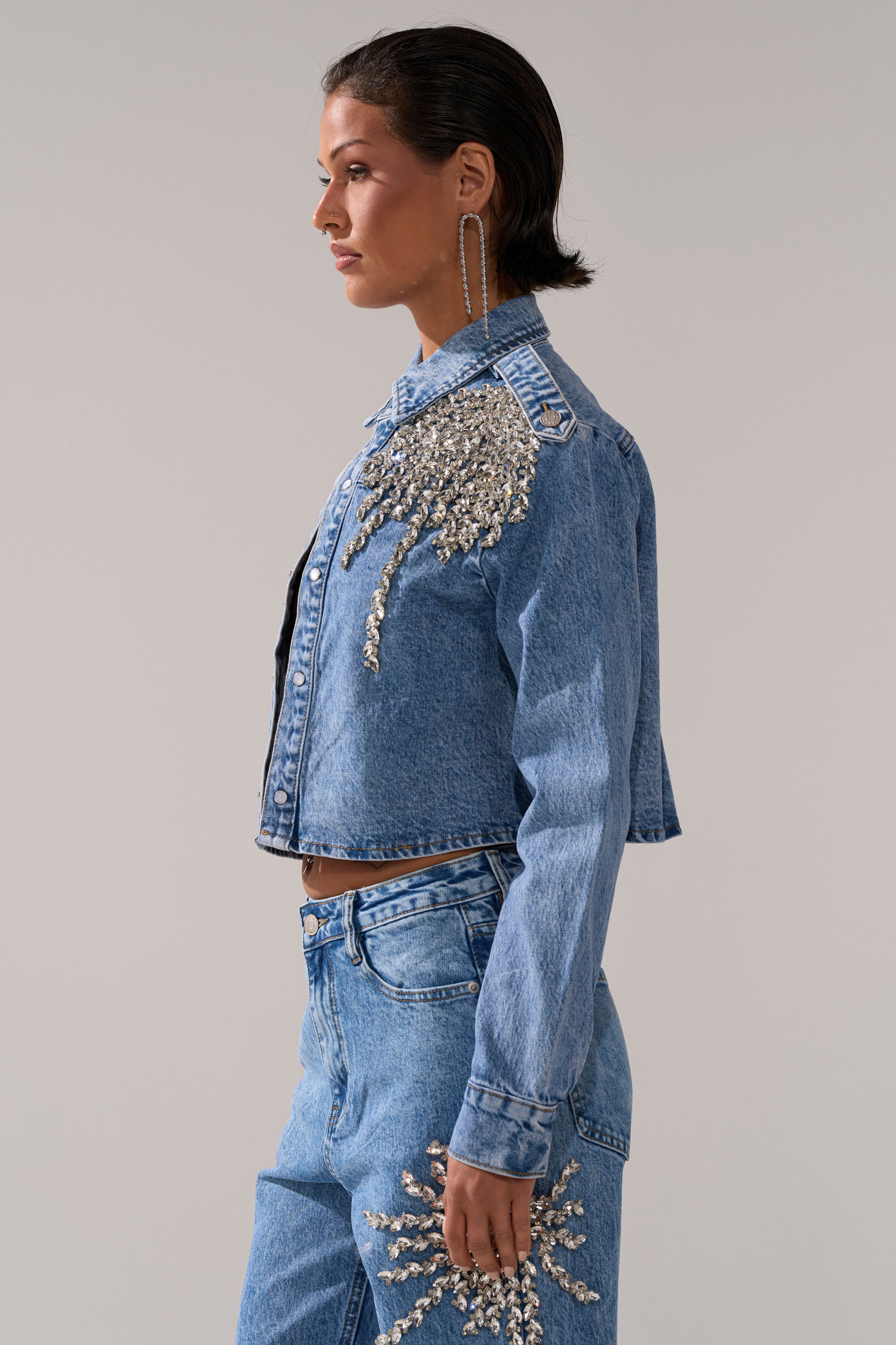 A woman with short dark hair and large hoop earrings models the SPARKLE AND SHINE QUEEN DENIM BUTTON DOWN, a cropped rhinestone jacket paired with embellished jeans, standing against a plain gray background.