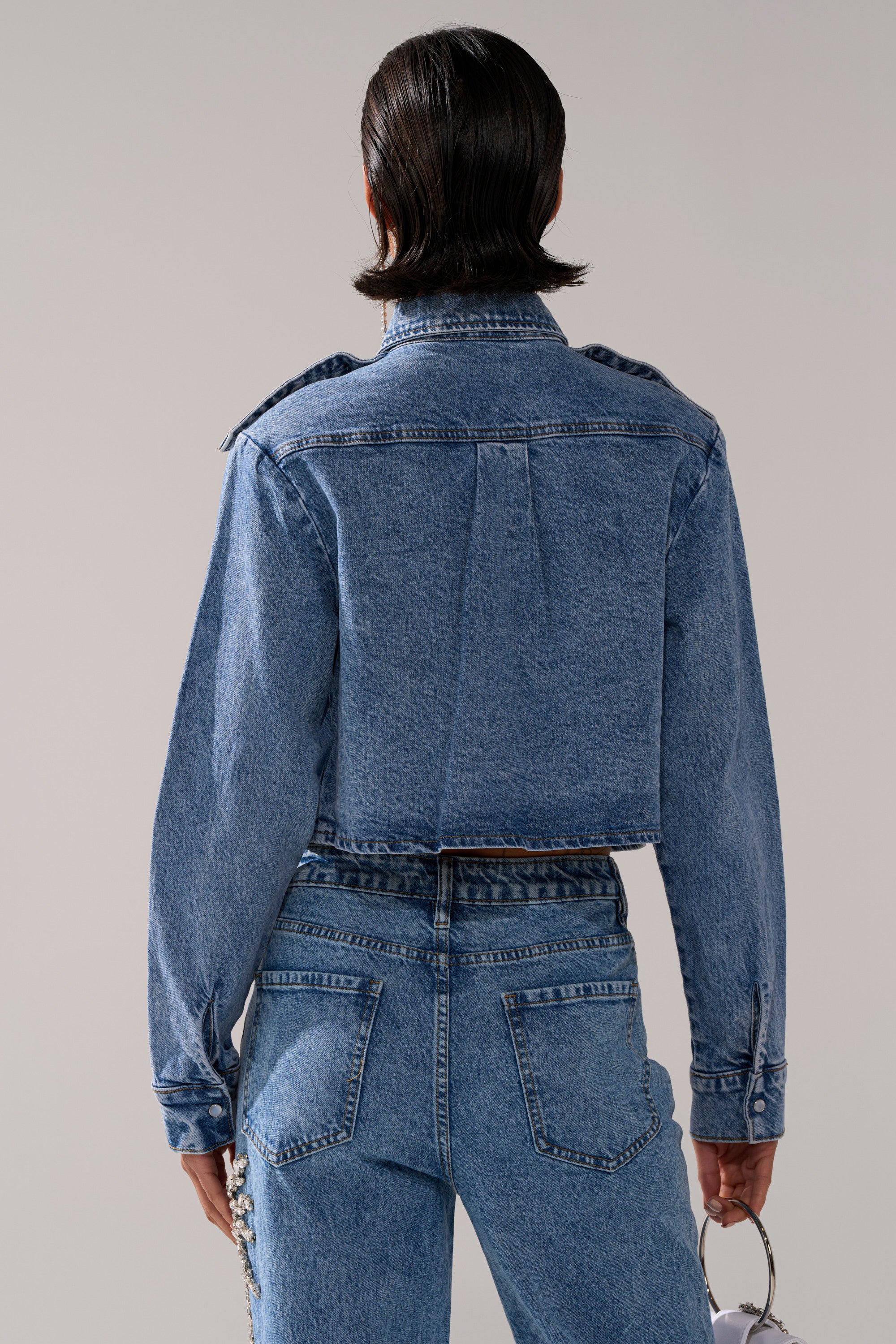 A person with short dark hair is shown from the back, wearing the SPARKLE AND SHINE QUEEN DENIM BUTTON DOWN and high-waisted blue jeans, holding sunglasses in their right hand against a plain neutral background.