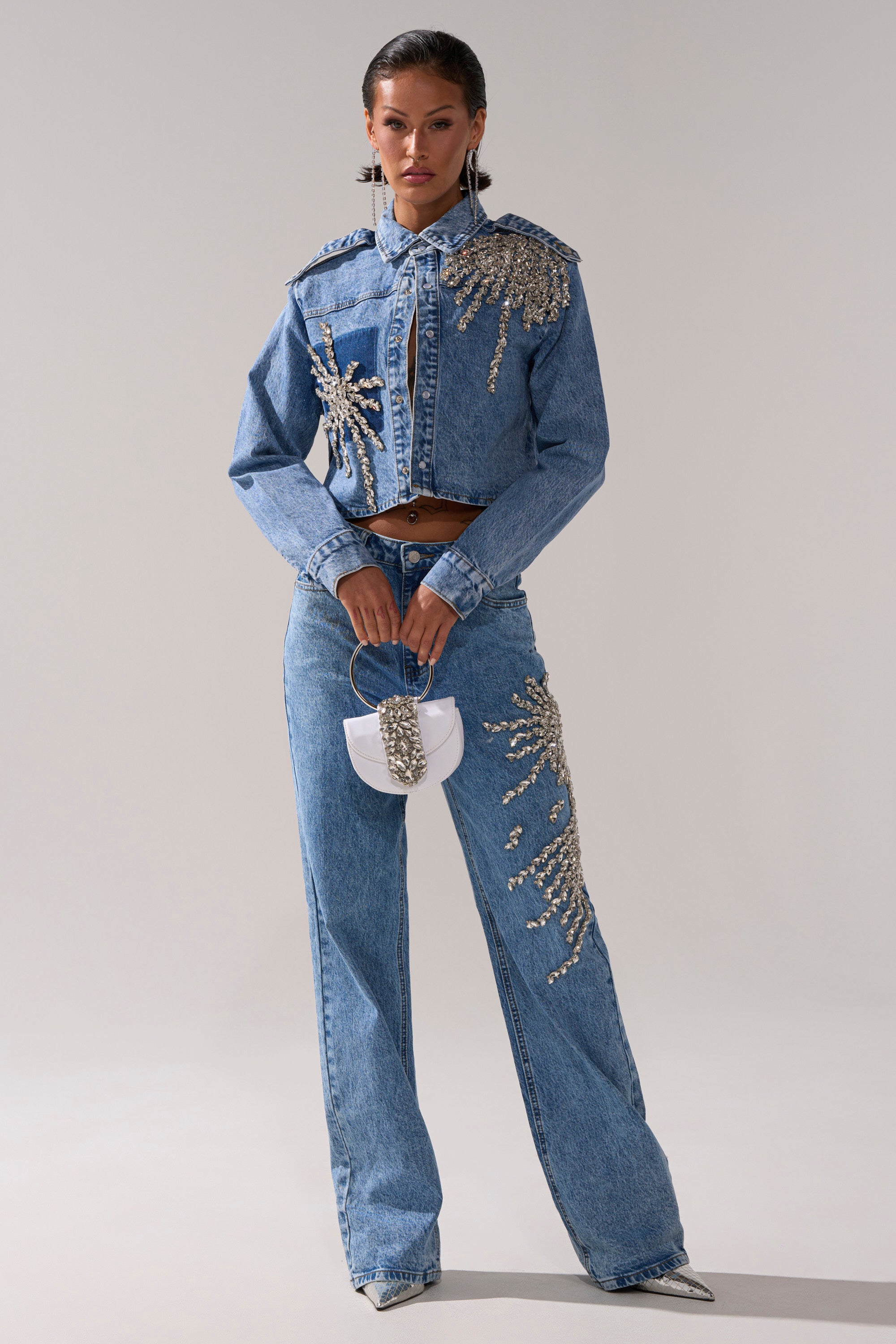 A woman stands confidently in the SPARKLE AND SHINE QUEEN DENIM BUTTON DOWN, a cropped, rhinestone-embellished jacket paired with high-waisted jeans, a small white purse with silver accents, slicked-back hair, and pointed-toe shoes.