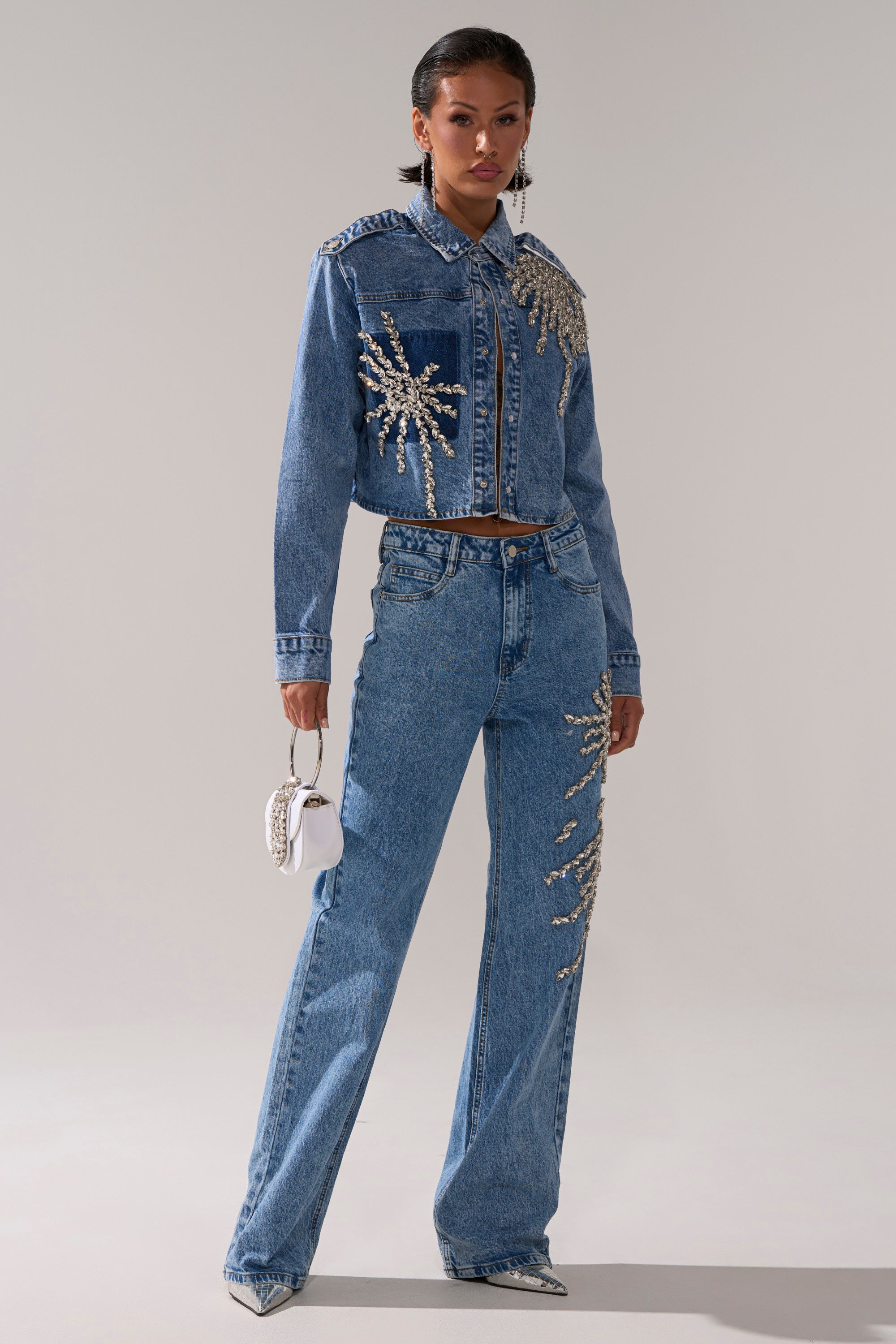 A woman stands confidently in the SPARKLE AND SHINE QUEEN DENIM BUTTON DOWN with matching jeans, both embellished with rhinestone designs. She accessorizes with a white handbag and pointed silver heels, set against a simple light background.