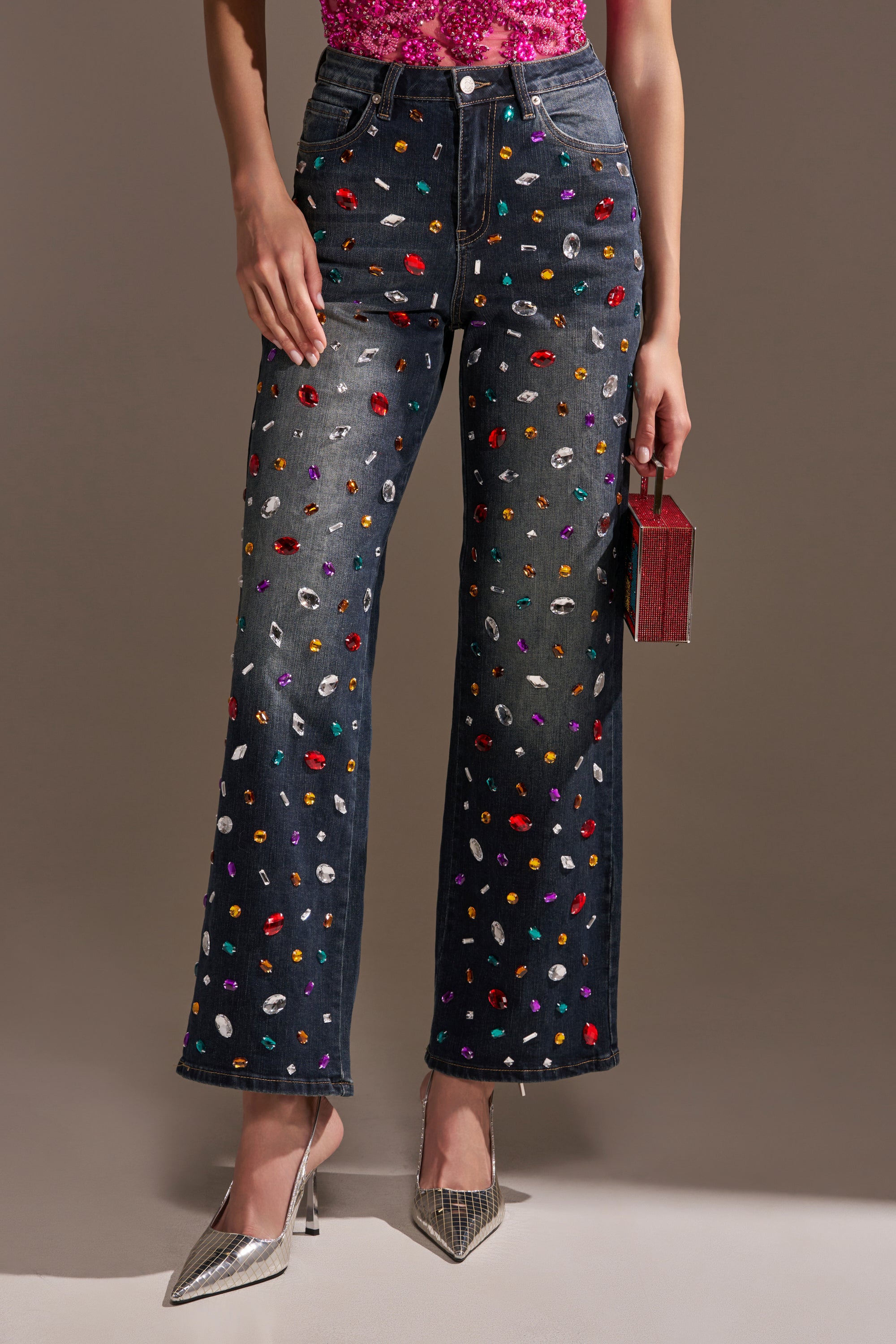 The lower half of a person in JEWELERS ROW RHINESTONE DENIM JEANS, covered with colorful gems, paired with a sparkly pink top, silver heels, and a small glittery purse is shown.