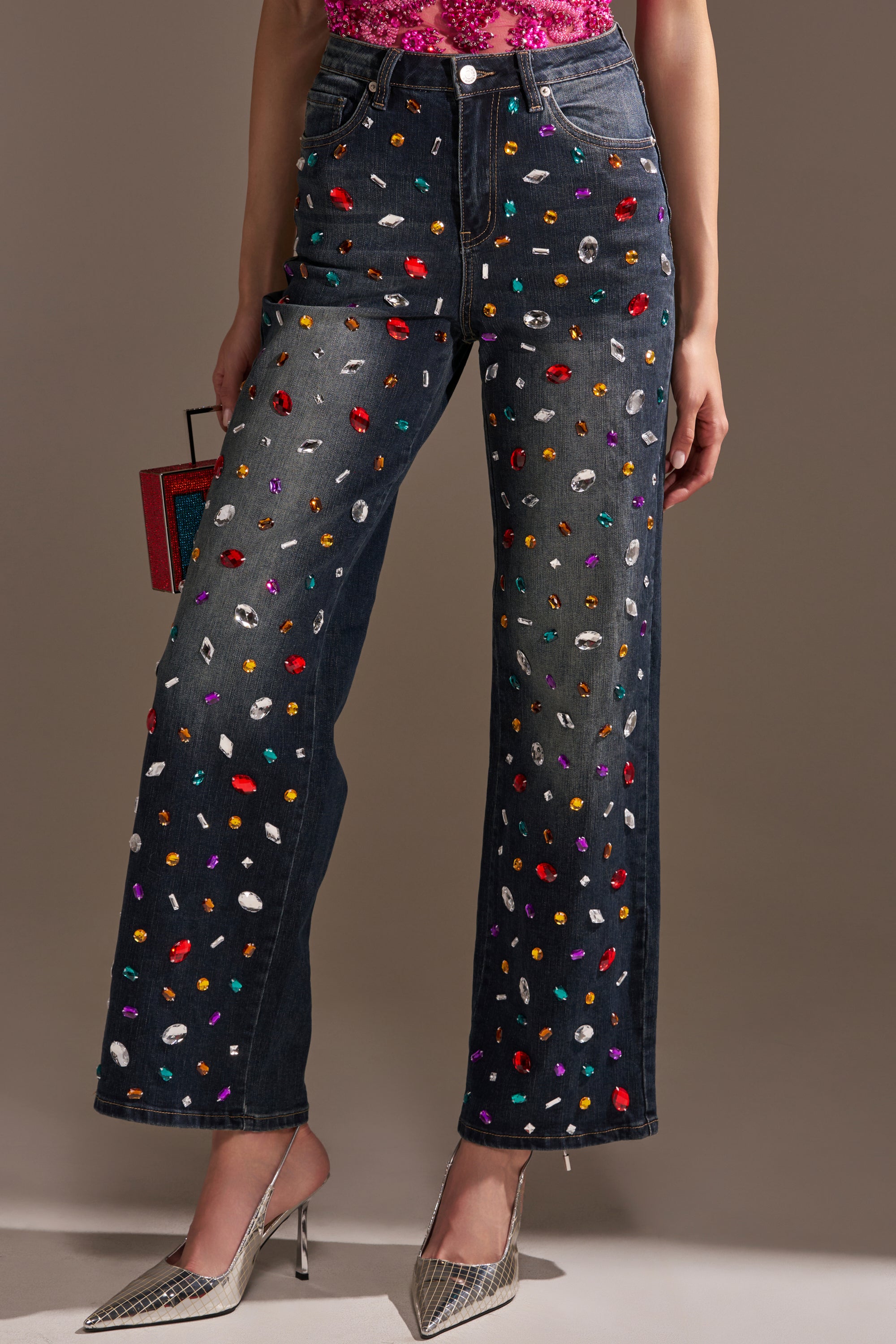 A person wears JEWELERS ROW RHINESTONE DENIM JEANS with colorful gemstone embellishments, a pink sequined top, silver pointed-toe heels, holds a small purse, and stands against a plain neutral background.