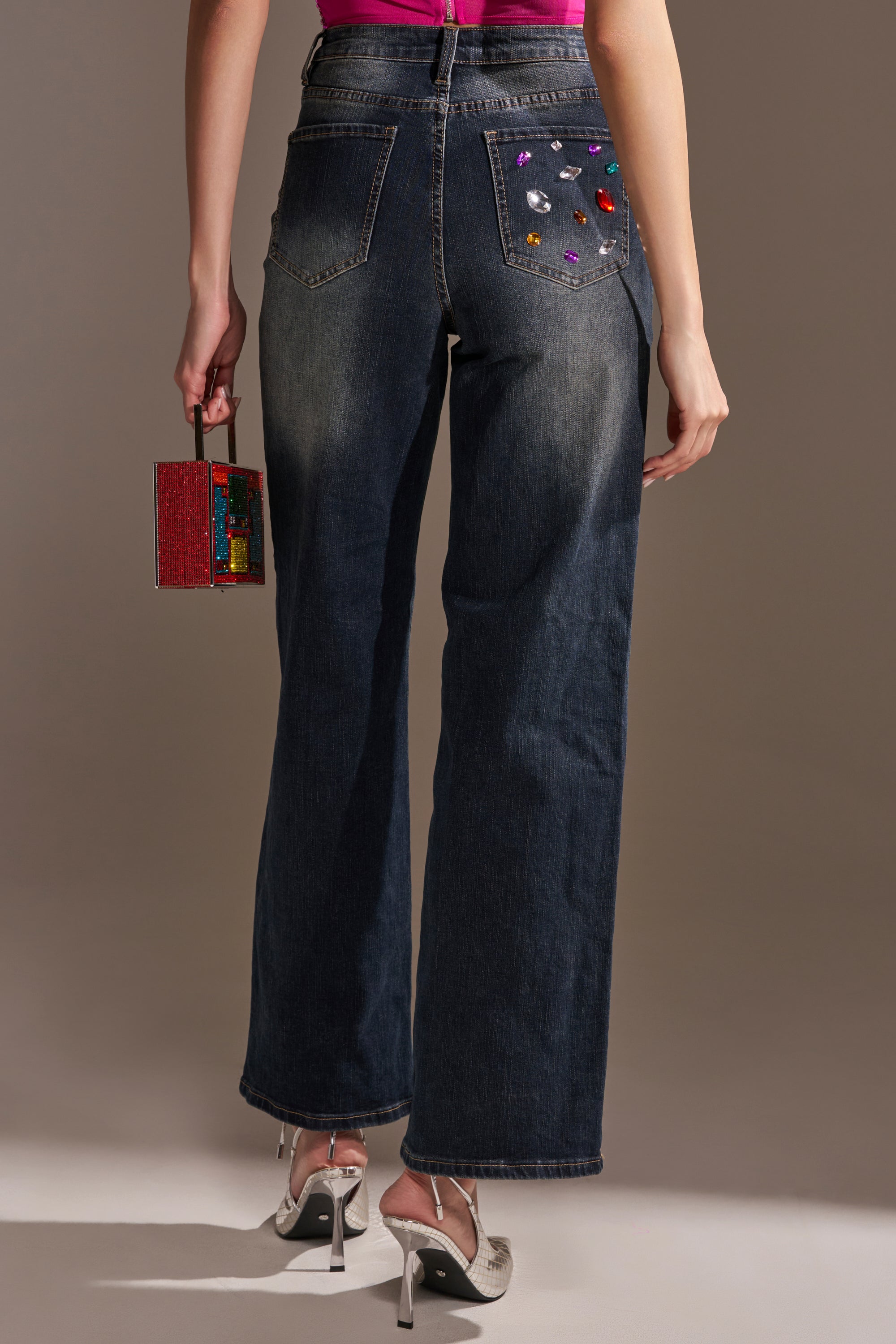 A person stands facing away, wearing JEWELERS ROW RHINESTONE DENIM JEANS with colorful gems on one back pocket, a pink top, silver heels, and holding a small red boxy handbag.