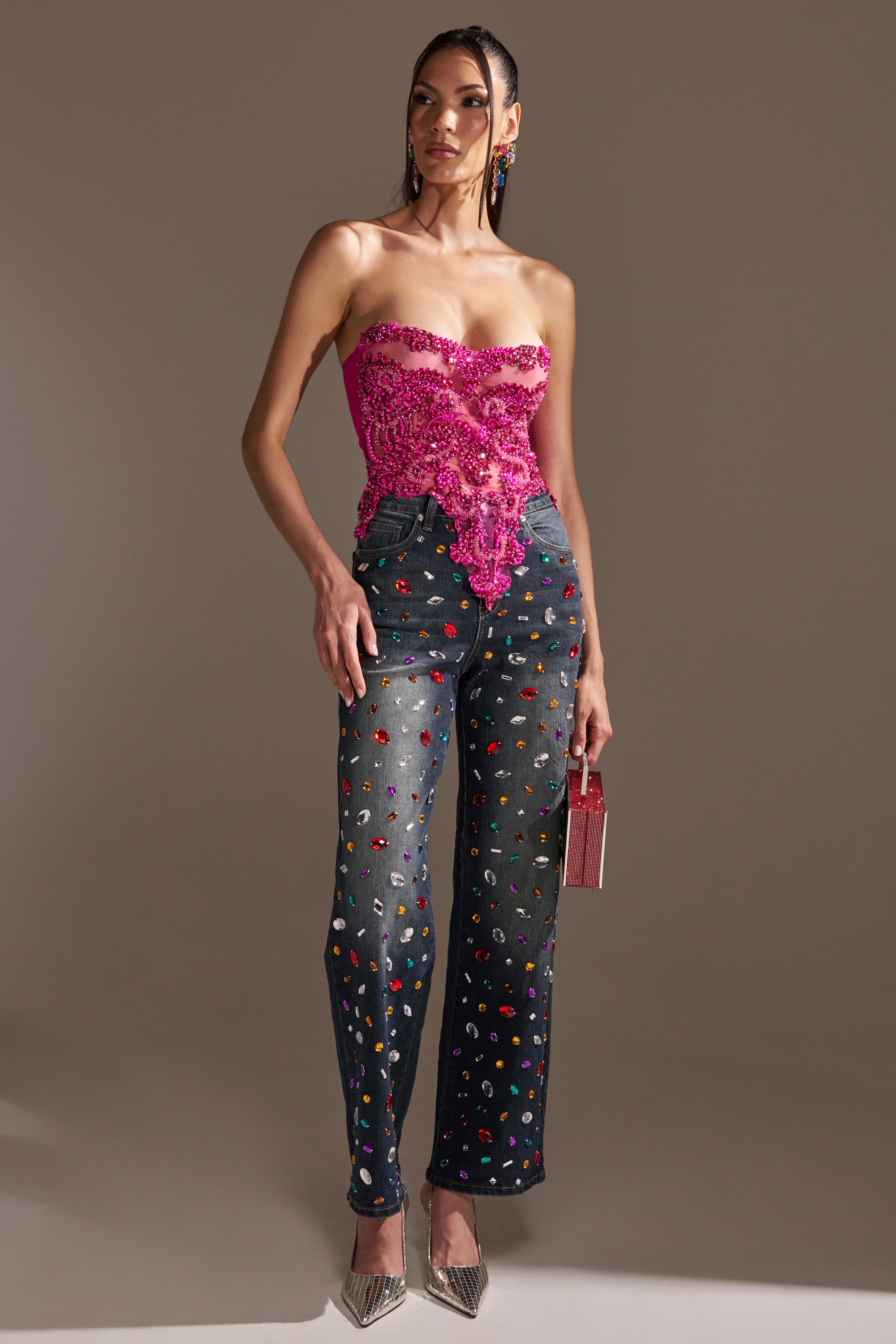 A woman poses in JEWELERS ROW RHINESTONE DENIM JEANS with a bright pink lace strapless top, silver pointed heels, and a small pink bag, creating a playful and sparkling look against a plain background.