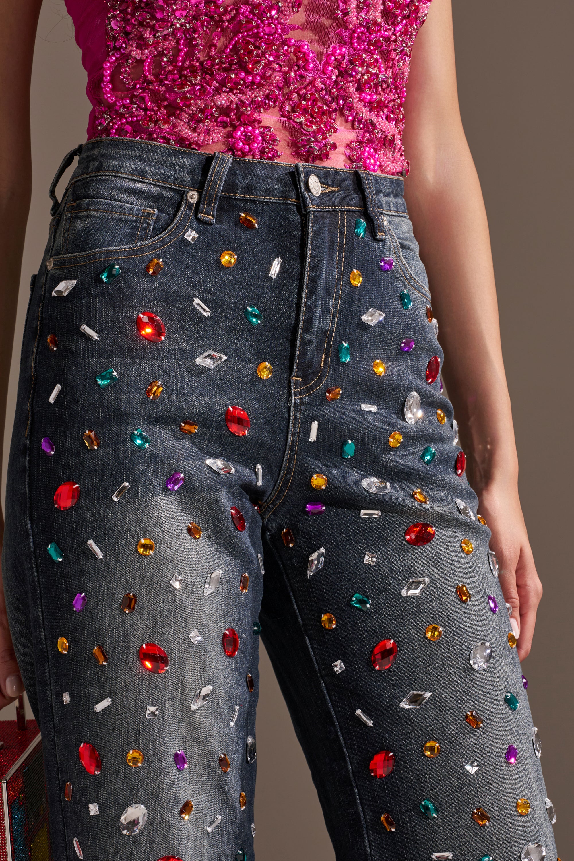 A person shown from waist to mid-thigh wears JEWELERS ROW RHINESTONE DENIM JEANS, adorned with colorful gems, paired with a bright pink beaded top.