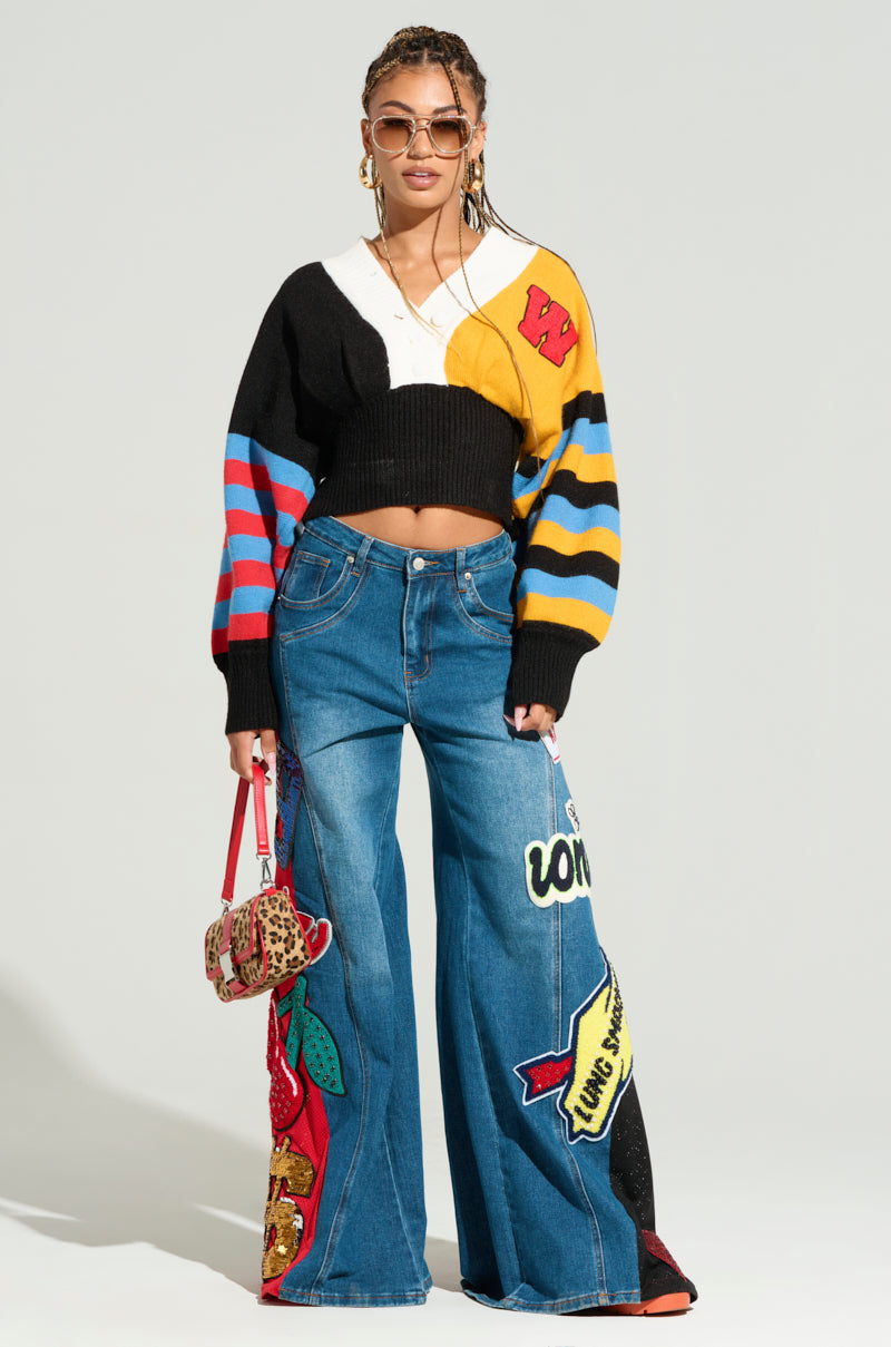 A woman wearing sunglasses, a multicolored knit sweater with various patterns, and the NOT SORRY PATCHWORK DENIM JEANS featuring embroidered patches. She holds a small leopard print handbag and stands against a plain background.