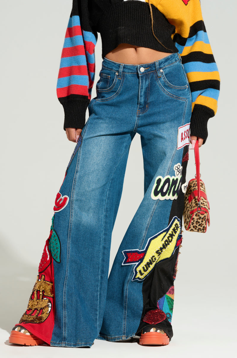 A person wearing NOT SORRY PATCHWORK DENIM JEANS paired with an embroidered black striped sweater, holding a small leopard print bag and wearing orange platform shoes. The outfit showcases bold, eclectic patterns.