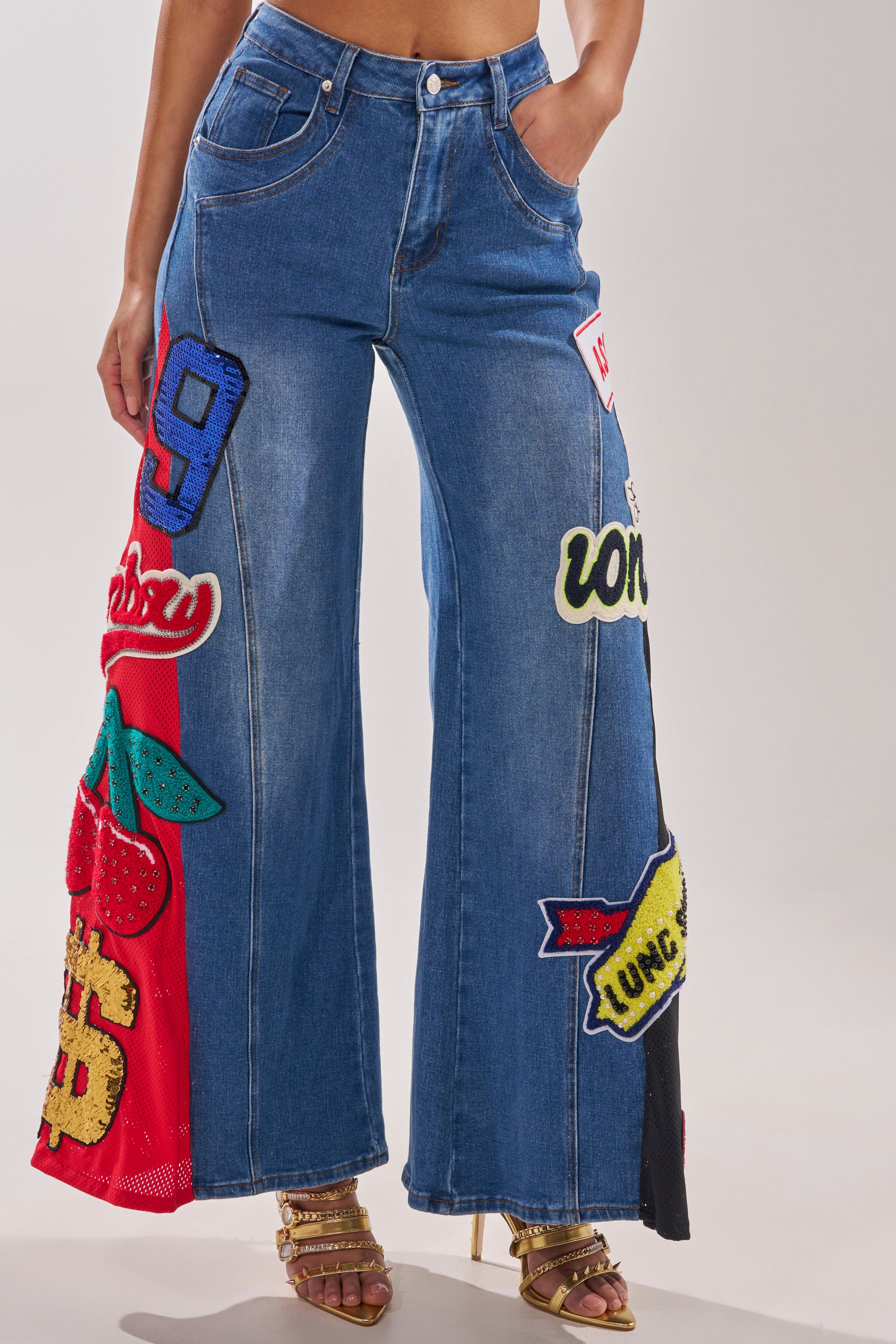 A person in NOT SORRY PATCHWORK DENIM JEANS with bold fabric patches—numbers, letters, a strawberry, and a dollar sign—styled with gold high-heeled sandals.