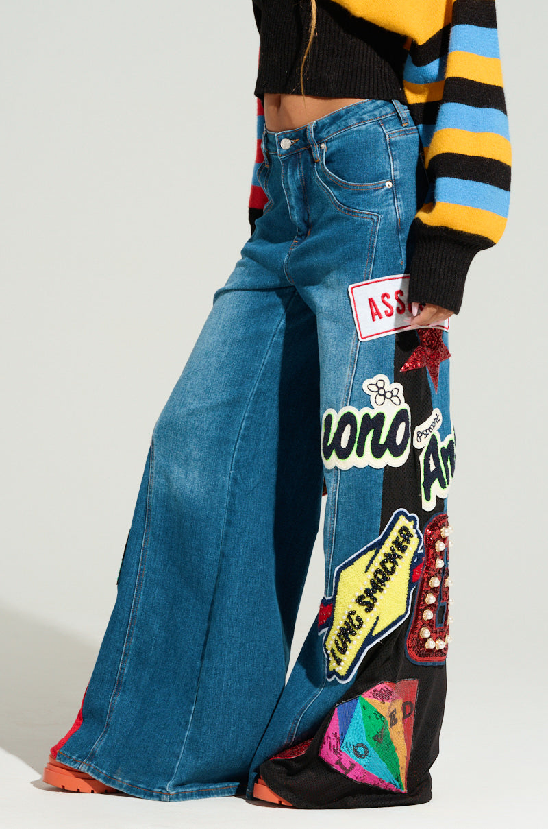 The image shows a person's lower torso and legs wearing the NOT SORRY PATCHWORK DENIM JEANS, featuring a high-waisted, wide-leg silhouette with blue denim decorated with various colorful embroidered patches and words. Completing the look is a long-sleeve sweater patterned in yellow, blue, and black horizontal stripes.