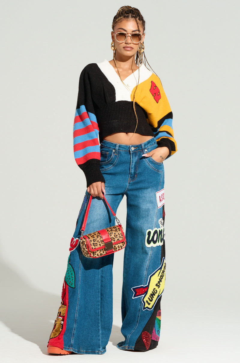 A woman in stylish attire poses confidently, flaunting her fashion-forward edge. She wears a black cropped sweater with multicolored sleeves, sunglasses, and hoop earrings. Her NOT SORRY PATCHWORK DENIM JEANS feature a wide-leg silhouette and vibrant embroidered patches. Holding a leopard-print handbag, she exudes boldness and trendiness.