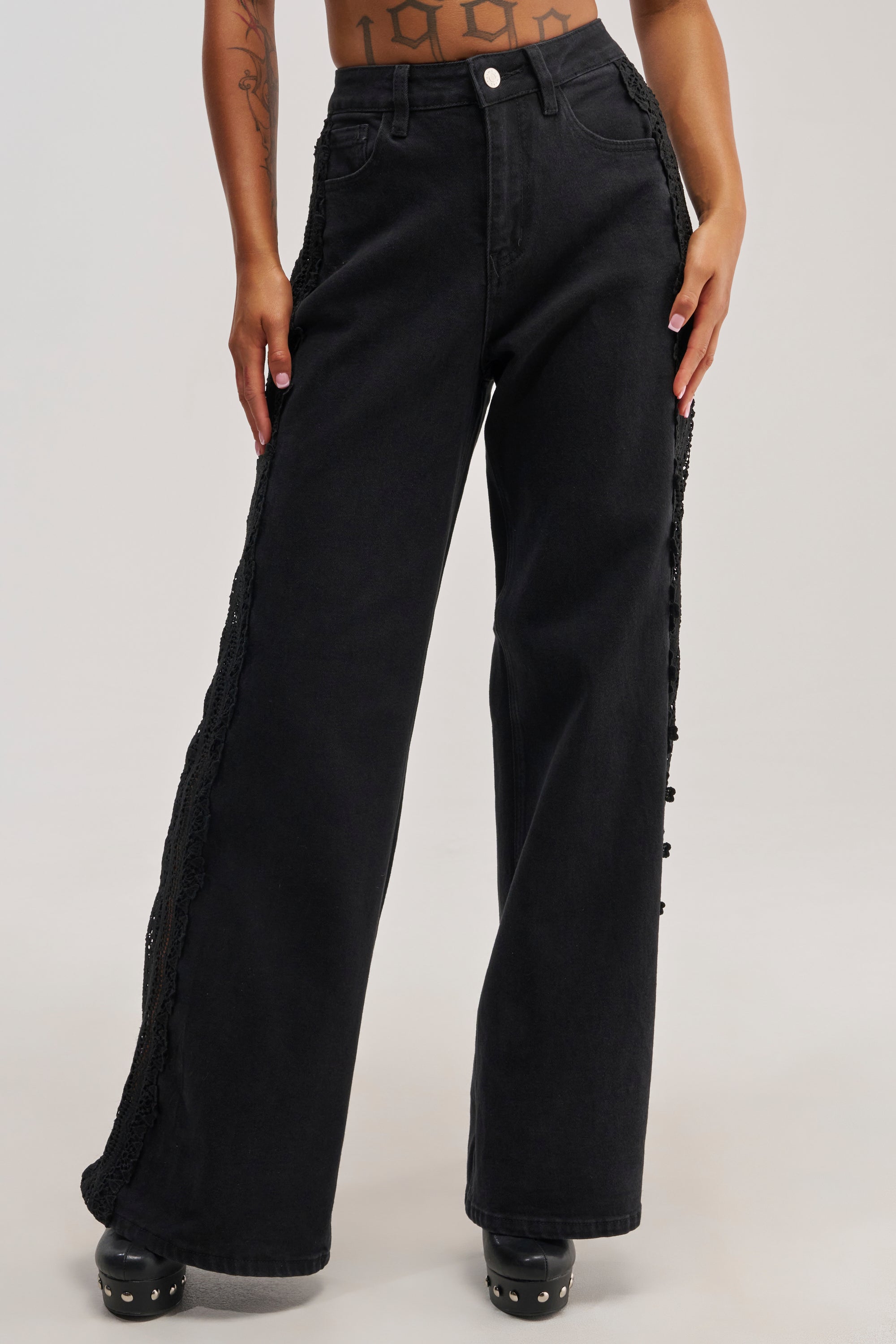 Wearing the LOCKED IN LACE DENIM—high-rise black wide-leg jeans featuring delicate lace trim along the sides—paired with black platform shoes and a cropped top, this person’s tattoos are visible on their midriff and arms.