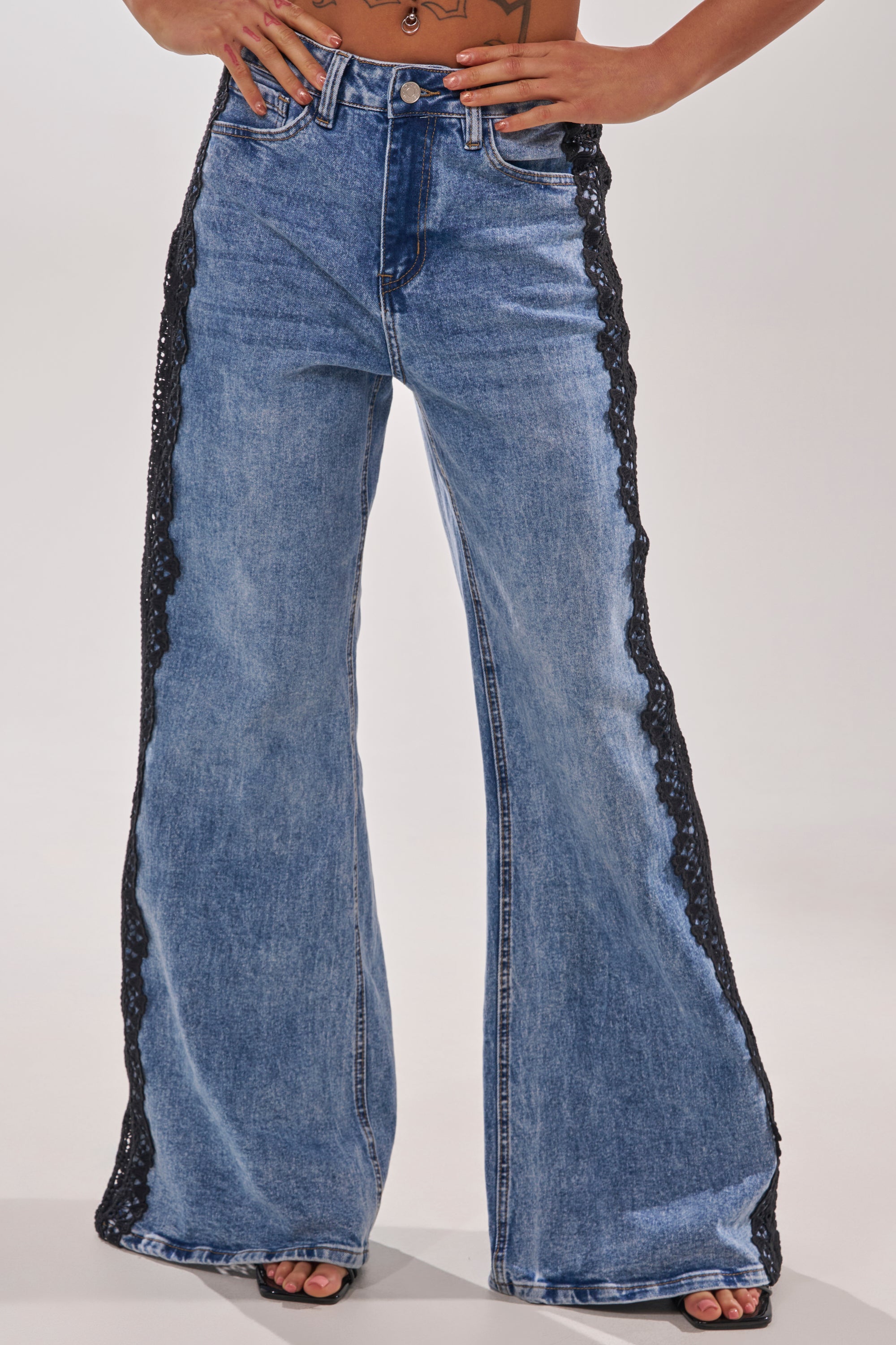 A person stands against a plain background, hands on hips, wearing LOCKED IN LACE DENIM—high-rise, wide-leg jeans with black lace trim down the outer sides, button closure, and pockets. 0