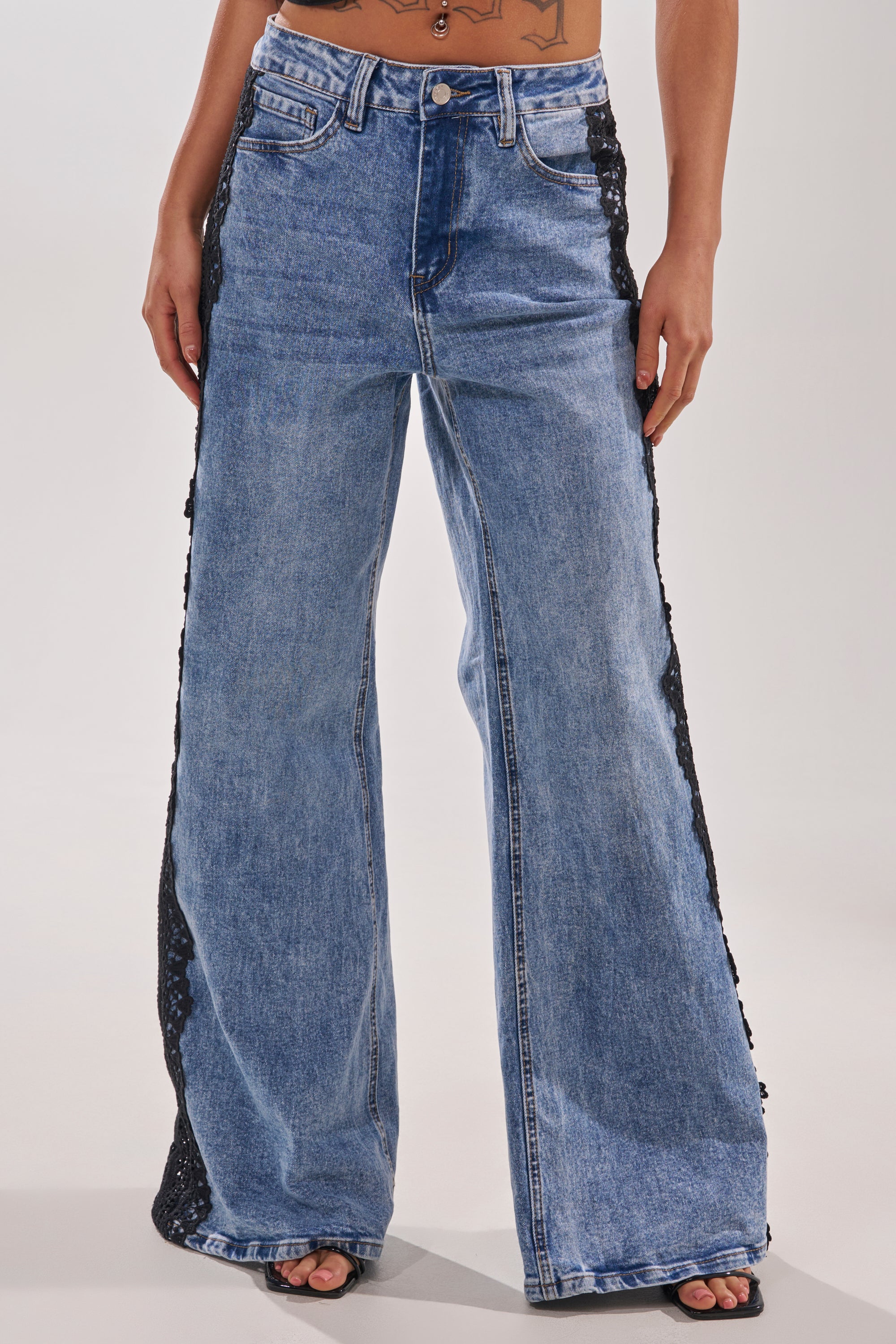 Person wearing LOCKED IN LACE DENIM, high-rise wide-leg jeans with black sequin panels along the sides; standing with legs slightly apart and midriff plus tattoo visible above the waistband. 1