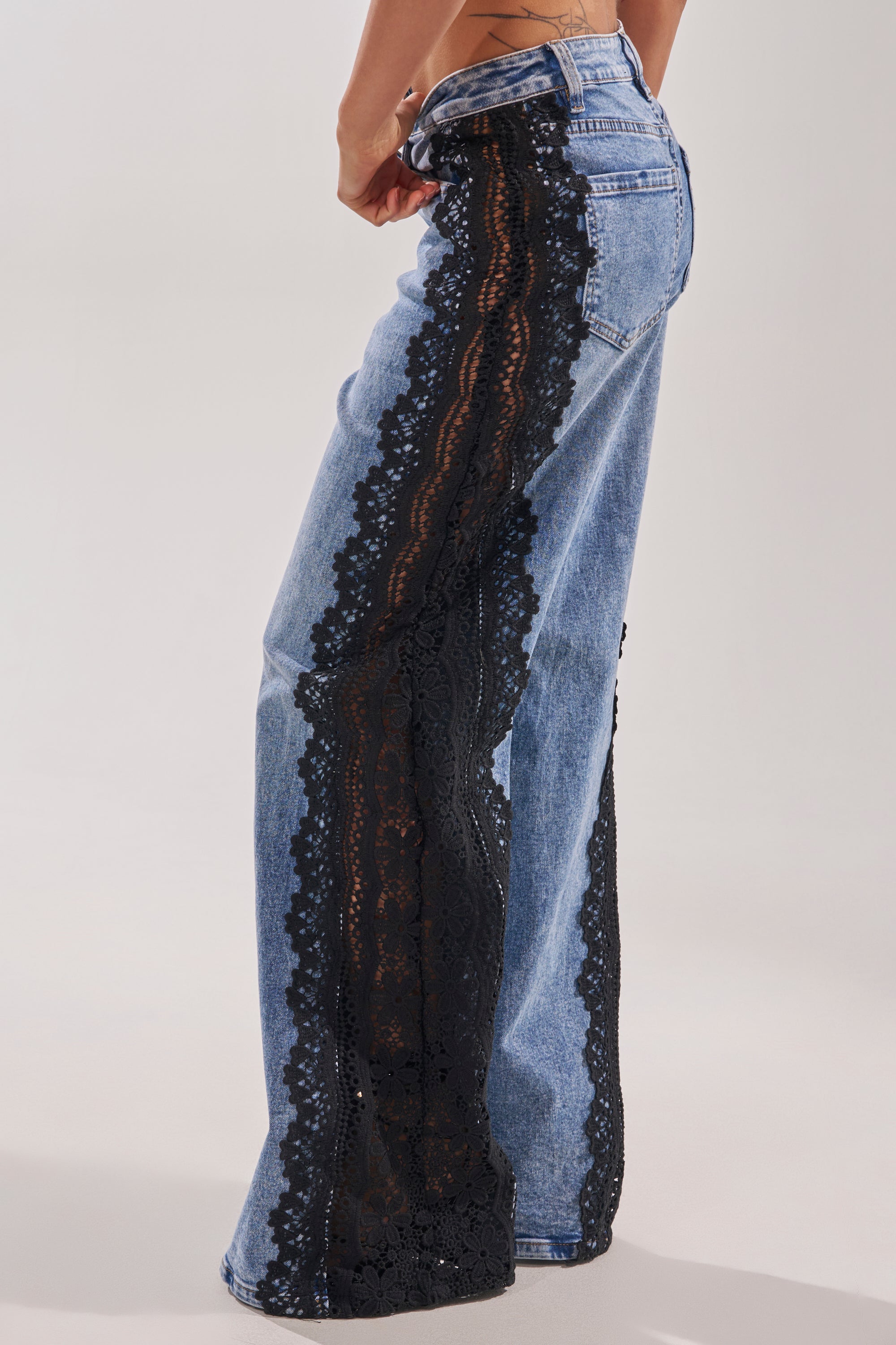 Someone models the LOCKED IN LACE DENIM, wide-leg, high-rise jeans featuring a bold black lace trim down the side seams, showcased from a side view against a plain light background. 2
