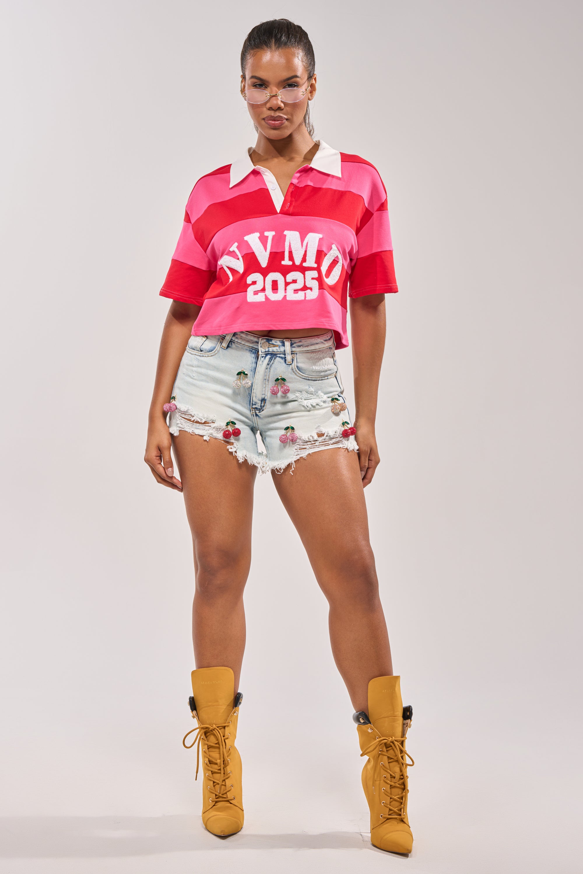 A woman faces forward wearing a pink and white collared shirt with "NVMO 2025" text, CHERRY DENIM SHORT, and tan lace-up boots against a plain light background. 5