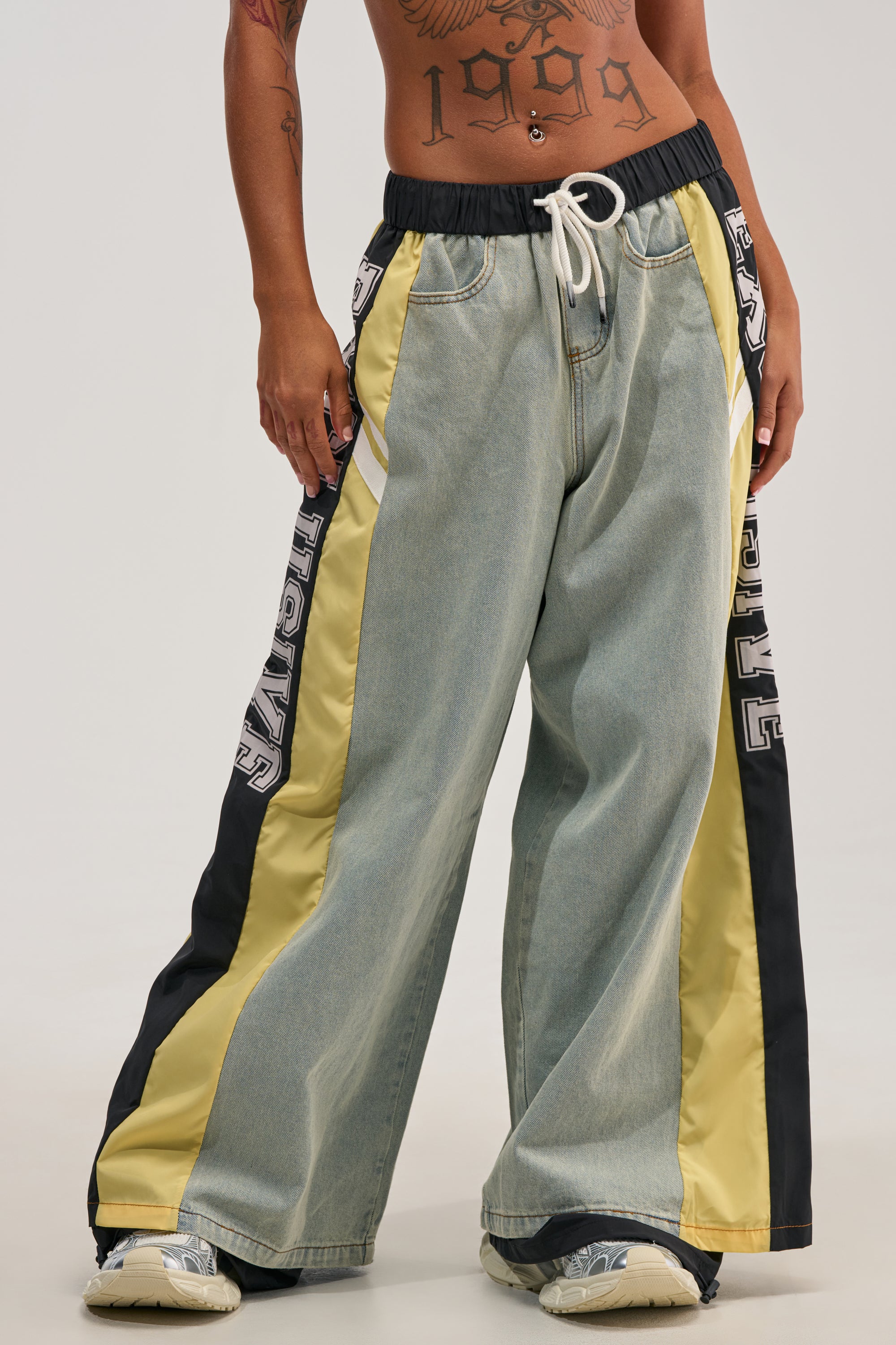 Wearing the EXCLUSIVE JOGGER, which features wide-leg streetwear styling with yellow and black side panels, bold black-and-white lettering, and a drawstring waist; the model's tattooed midriff is visible along with chunky white sneakers. Only the lower body is shown.
