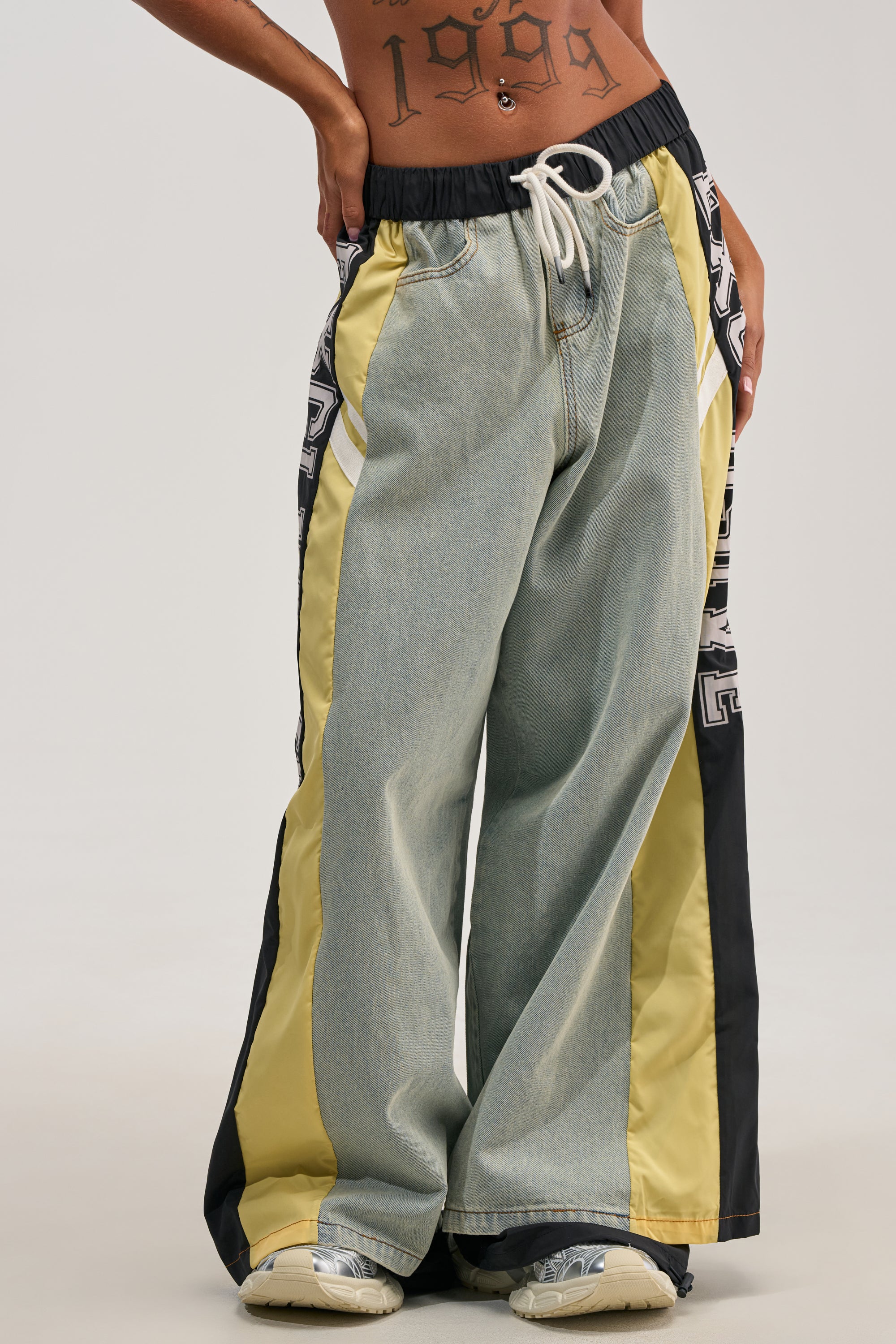 Wearing the EXCLUSIVE JOGGER, which features loose-fitting denim with striking yellow and black side panels and a comfortable drawstring waist, paired with off-white sneakers. An abstract tattoo peeks out from their lower stomach, adding to the laid-back streetwear vibe.