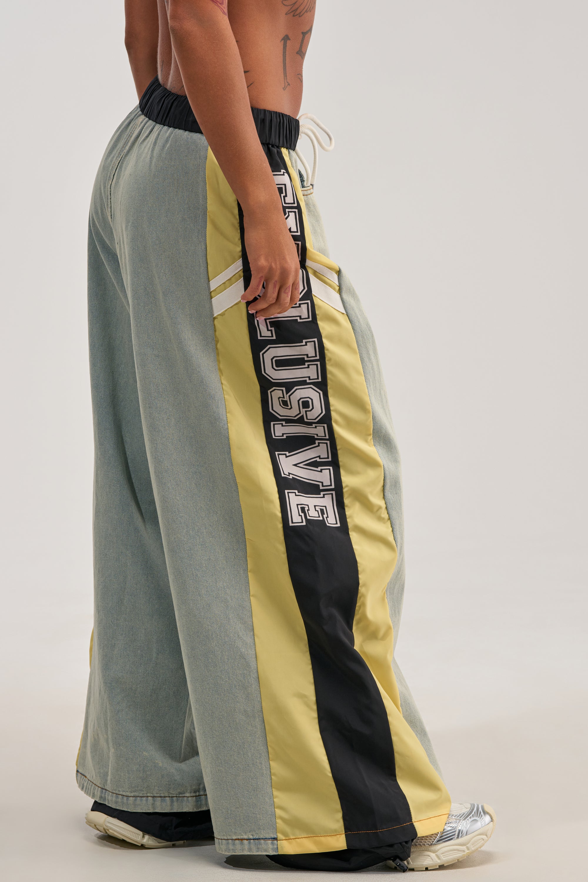 A lower torso and legs model the EXCLUSIVE JOGGER—loose, wide-leg denim joggers featuring light blue denim on the back and a striking yellow, black, and cream side panel emblazoned with bold “EXCLUSIVE” lettering for a standout streetwear look.