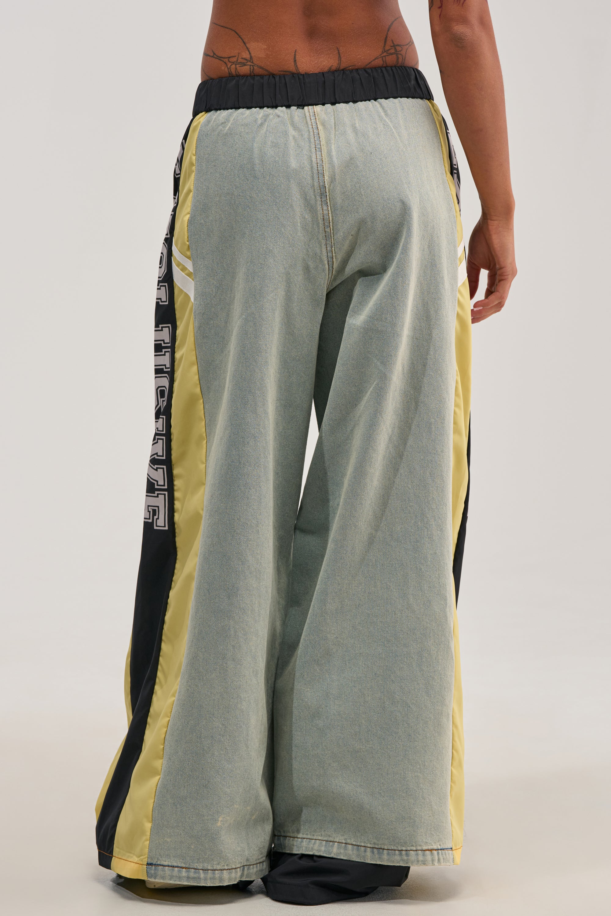 A person is shown from the back wearing the EXCLUSIVE JOGGER—loose, wide-leg light denim joggers featuring striking yellow and black side panels and a black elastic waistband, creating a bold streetwear look.