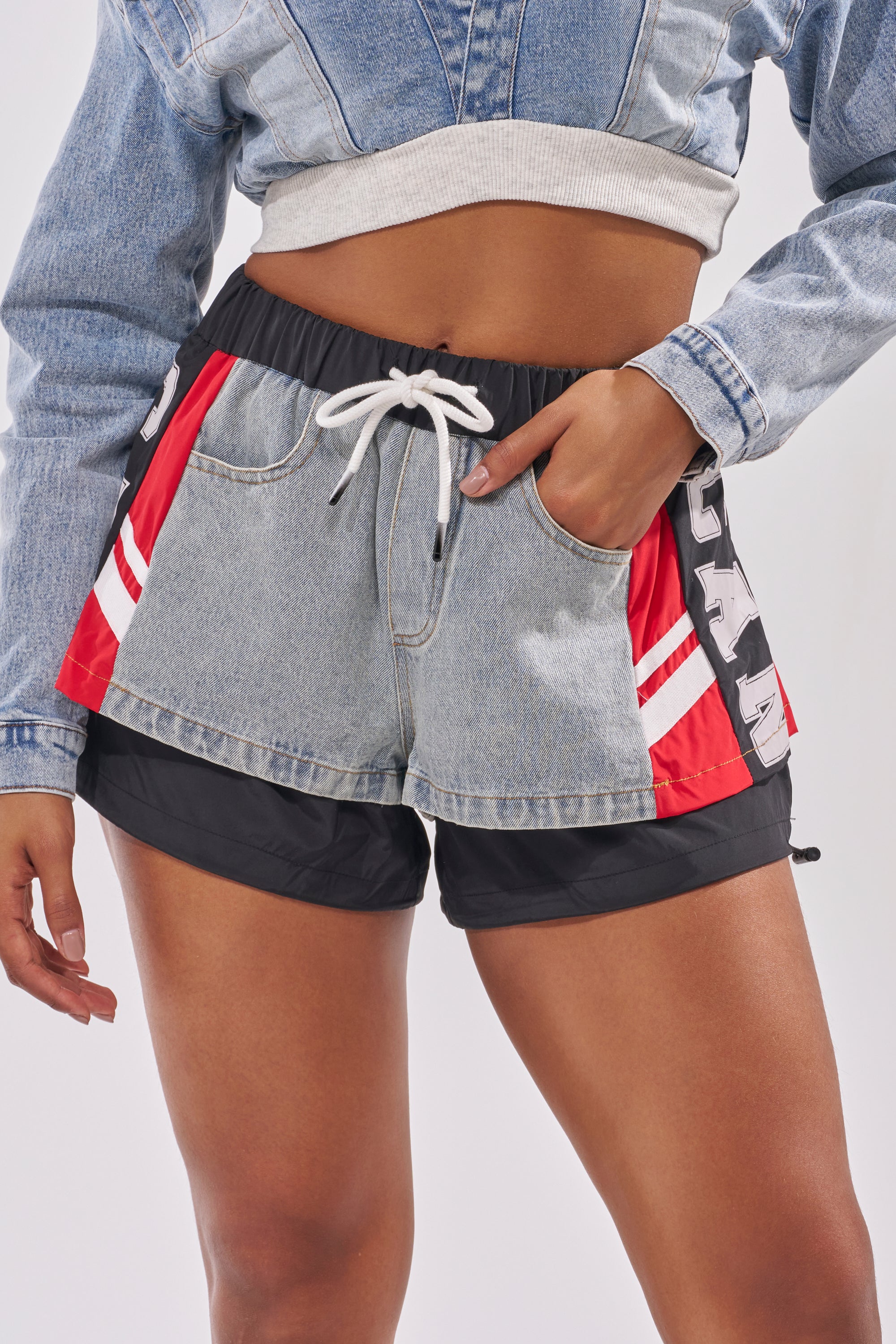 A person wears the LETS WIN SHORT—layered, denim color block shorts with red and white stripes over black athletic shorts, paired with a cropped denim jacket and finished with a white drawstring at the waistband for sporty streetwear style. 1