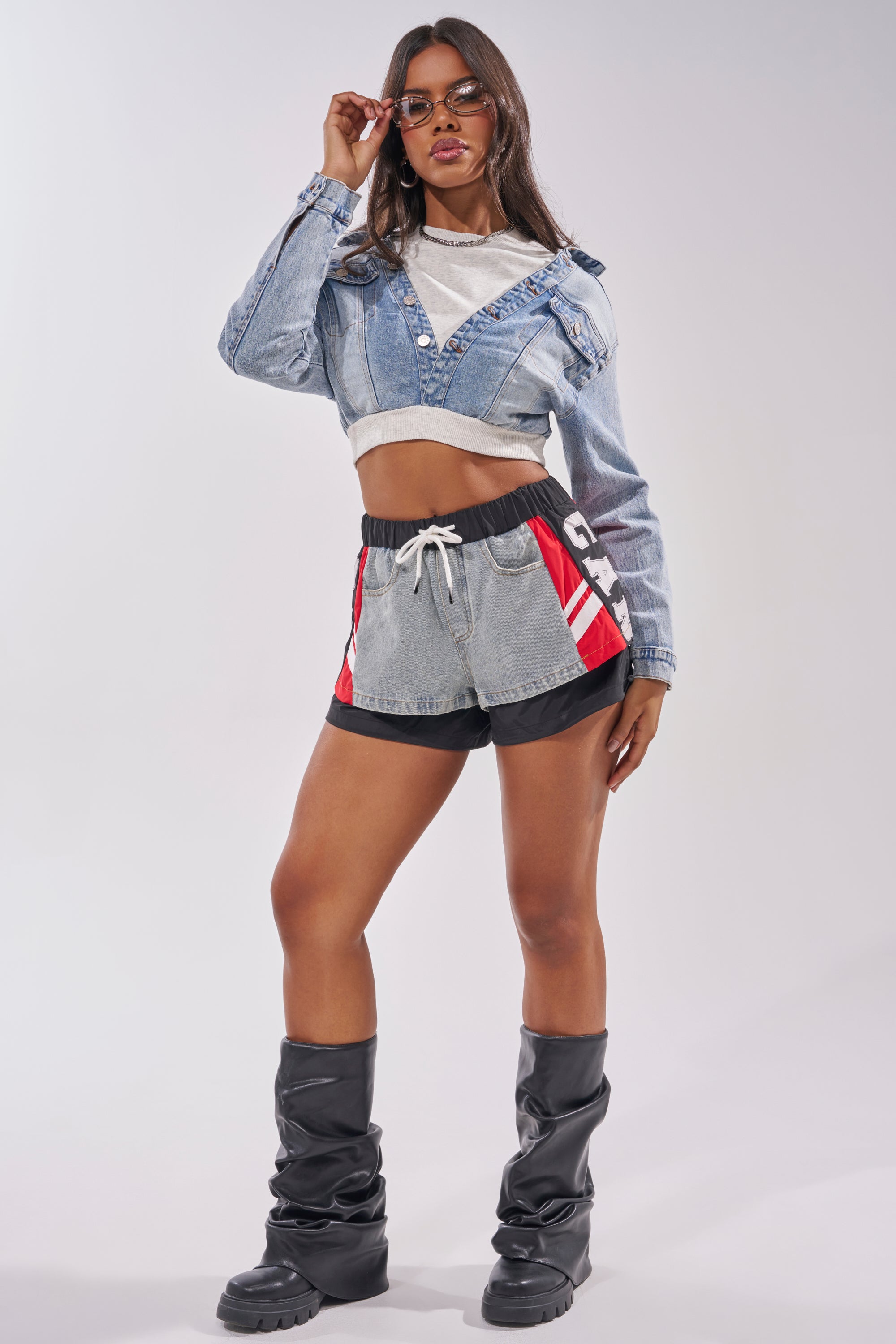 A woman poses confidently in sunglasses, a cropped denim jacket over a white top, LETS WIN SHORT retro-sport shorts with red and black accents, and tall black boots with thick soles. 8