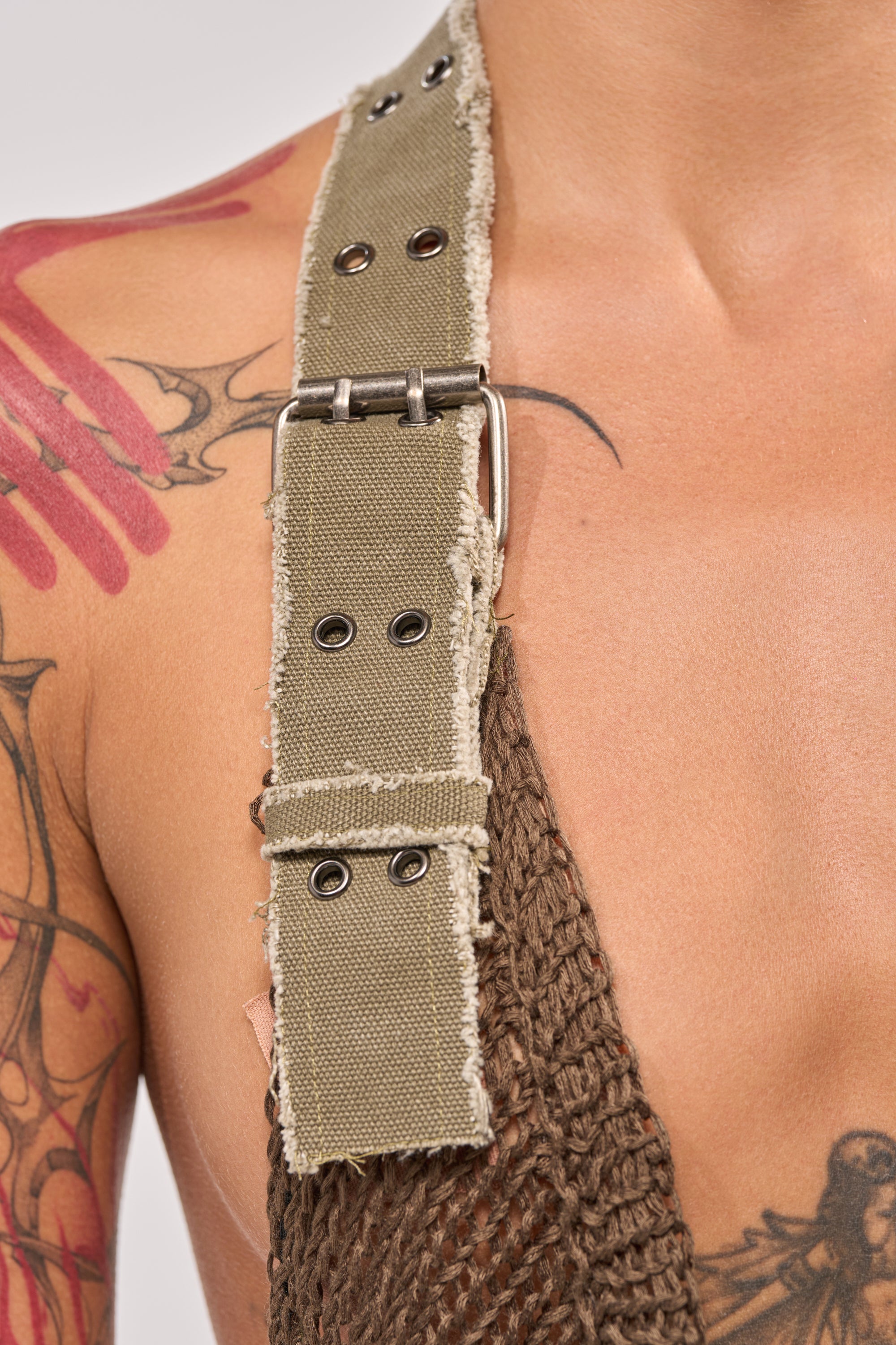 A close-up of a person's shoulder and upper chest shows tattoos, a large khaki strap with metal eyelets, and the MAKE A MESS KNIT MINI DRESS IN OLIVE—perfect for adding texture to your next festival dress look. 8