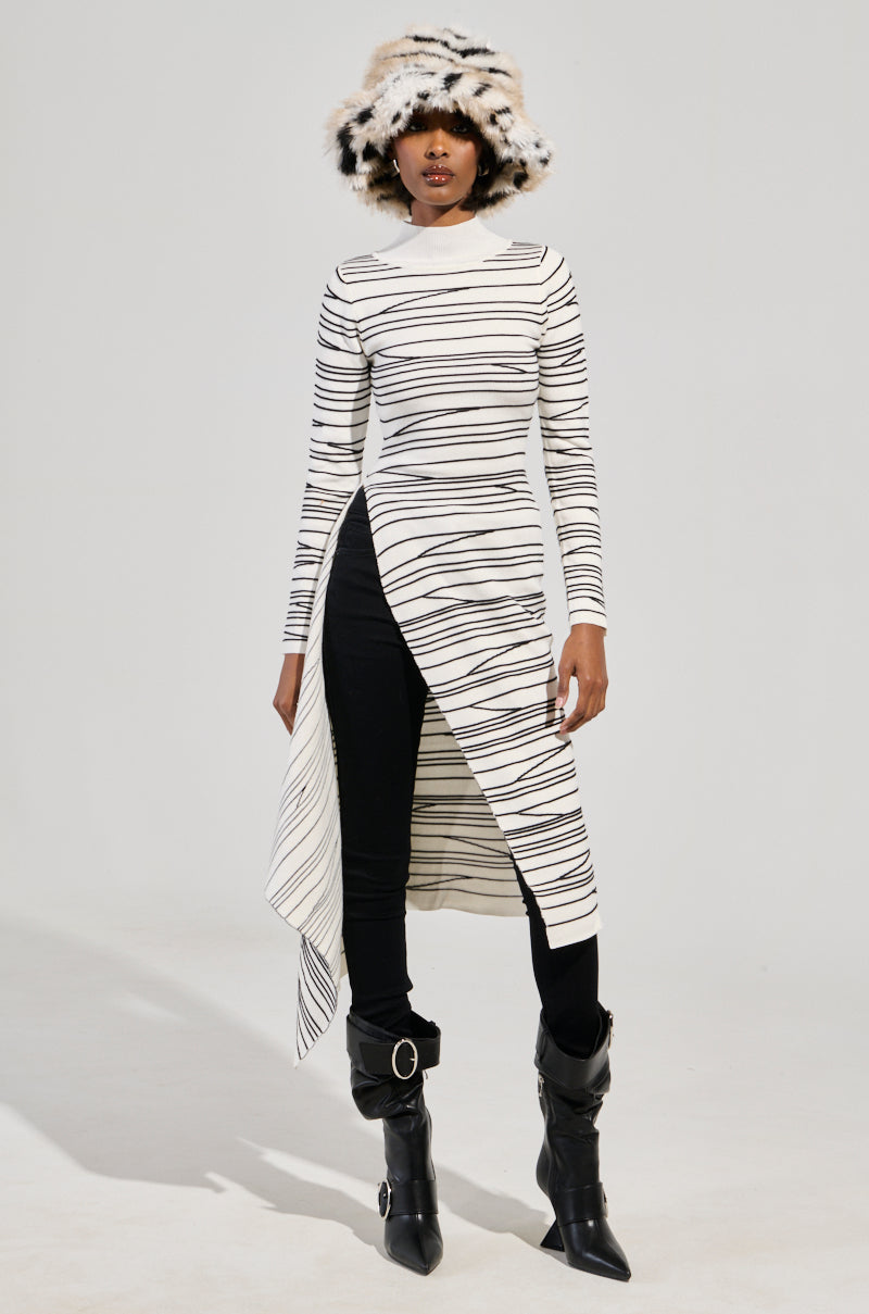 A person wearing the ISA High Low Sexy Slit Striped Sweater in white, styled as a dress with an asymmetrical hem, paired with black pants and buckle-adorned boots. They complete the look with a furry, patterned hat against a plain light gray backdrop.