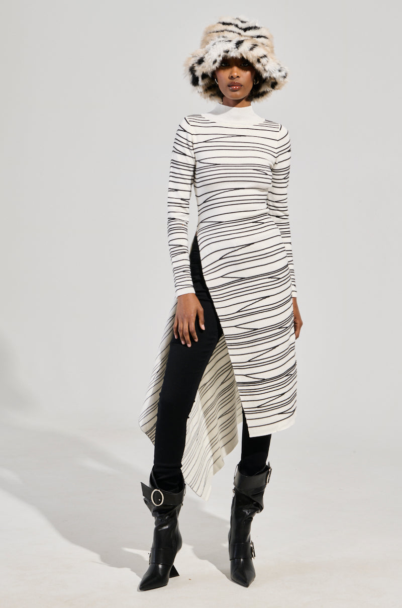 A person is dressed in the ISA High Low Sexy Slit Striped Sweater in white, paired with black pants and sleek black boots. They top off their look with a unique hat that showcases a mix of black and white fur patterns, creating a modern and fashionable ensemble against a plain background.