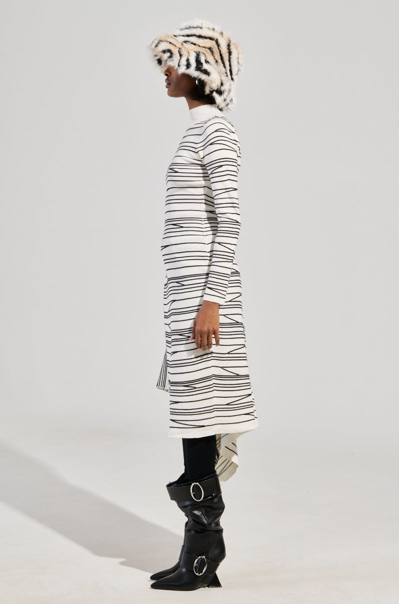 A person stands in profile wearing a zebra-patterned hat and the ISA High Low Sexy Slit Striped Sweater in white. The outfit is completed with black leggings and knee-high black boots featuring large circular buckles, adding a chic twist to the ensemble. The background is a plain, light-colored backdrop.