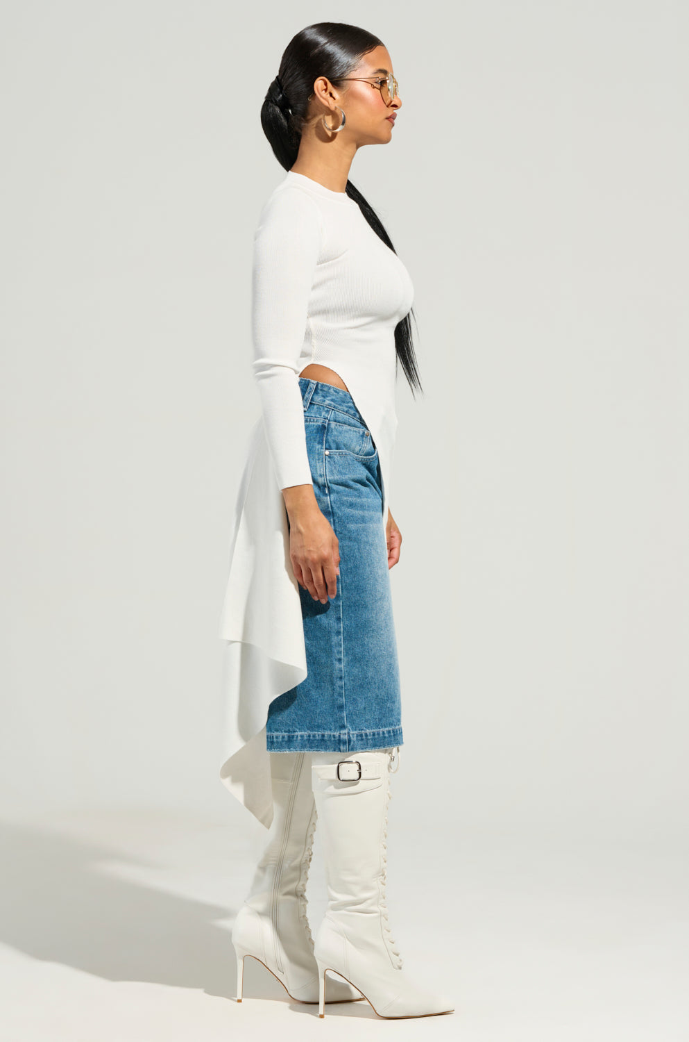 A woman with long black hair styled in a sleek ponytail is seen in profile, wearing glasses, the ISA HIGH LOW SEXY SLIT SWEATER in white, a blue denim skirt, and knee-high white lace-up heeled boots. She poses against a plain studio backdrop.