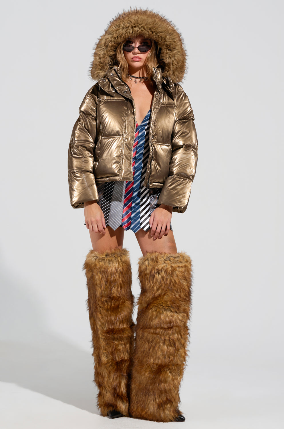 A person wearing the CAPRI FUR TRIM PUFFER jacket in a metallic polyester bronze style with an oversized hood, paired with large sunglasses, a plaid dress, and tall brown furry boots. The setting is a simple white studio backdrop.