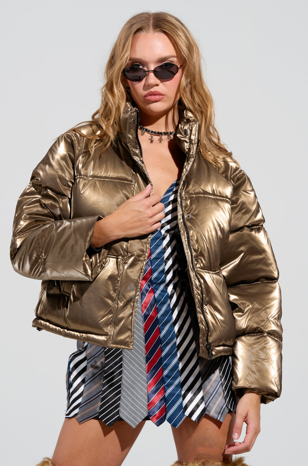 A woman with long hair showcases the CAPRI FUR TRIM PUFFER, a shiny metallic polyester jacket, paired with a dress crafted from assorted neckties. She completes her look with slim sunglasses and a choker necklace, while the oversized hood adds flair against a plain light background.