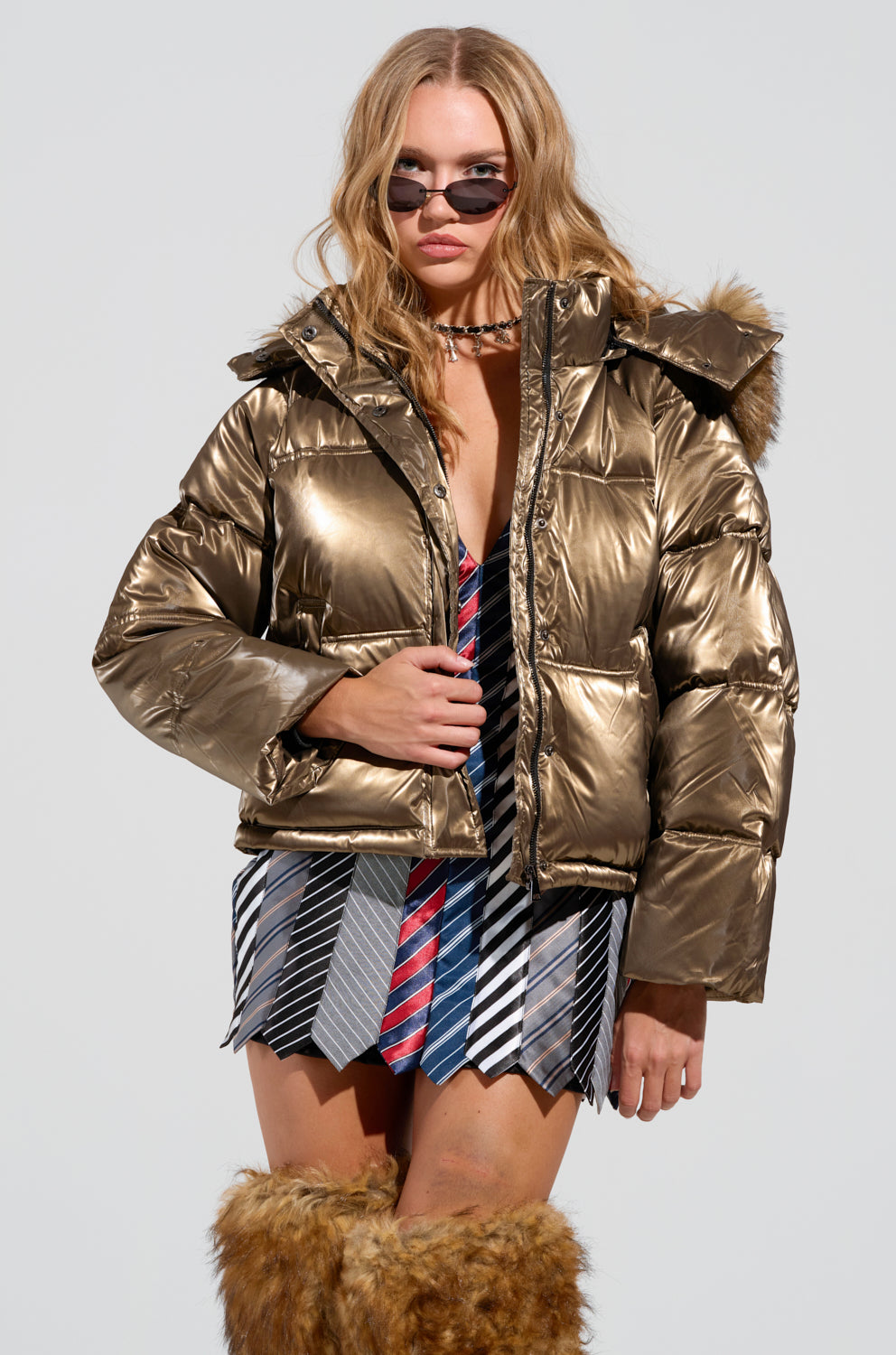 A woman stands confidently in the CAPRI FUR TRIM PUFFER jacket, its metallic polyester glistening under the light. She's wearing sunglasses, a short striped skirt, and furry knee-high boots. Her long hair flows freely as she poses against a plain background, effortlessly exuding style.