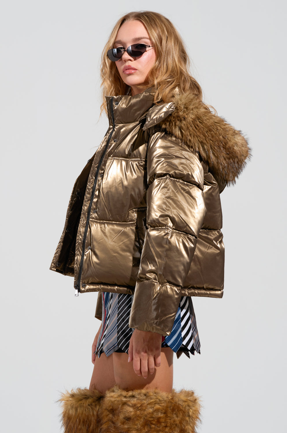 Dressed in a reflective, metallic gold CAPRI FUR TRIM PUFFER jacket with an oversized hood, accessorized with dark sunglasses, a striped skirt, and fur-covered boots, the individual strikes a confident pose against a simple backdrop.