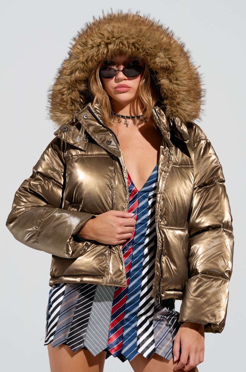 Against a plain backdrop, an individual is seen wearing the CAPRI FUR TRIM PUFFER, crafted from shiny metallic polyester. This jacket features a generous hood and luxurious fur lining. Complimented with sunglasses and a dress created from assorted neckties, they exhibit a distinctly unique fashion sense.