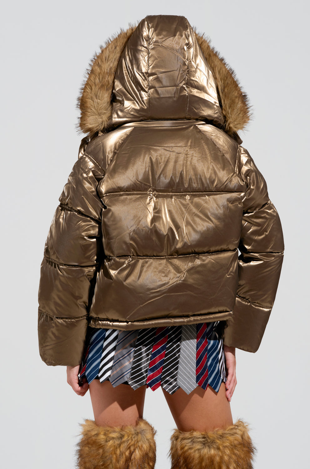 Wearing the striking Capri Fur Trim Puffer, a person in a glittering bronze jacket made of metallic polyester with an oversized hood faces away from the camera. Their outfit is completed with a skirt crafted from ties and fuzzy leg warmers, perfectly capturing the distinctive style of this look.
