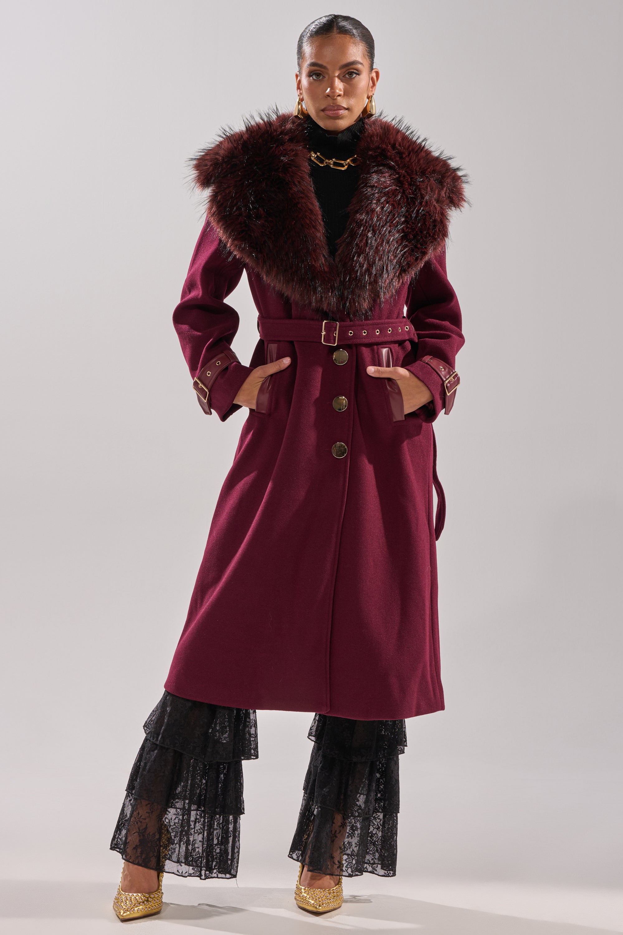Wearing the CATALINA FUR COLLAR TRENCH in burgundy with a large fur collar, gold buttons, and a belt, she puts her hands in the pockets, styled with black lace pants and gold shoes against a light grey background. 0