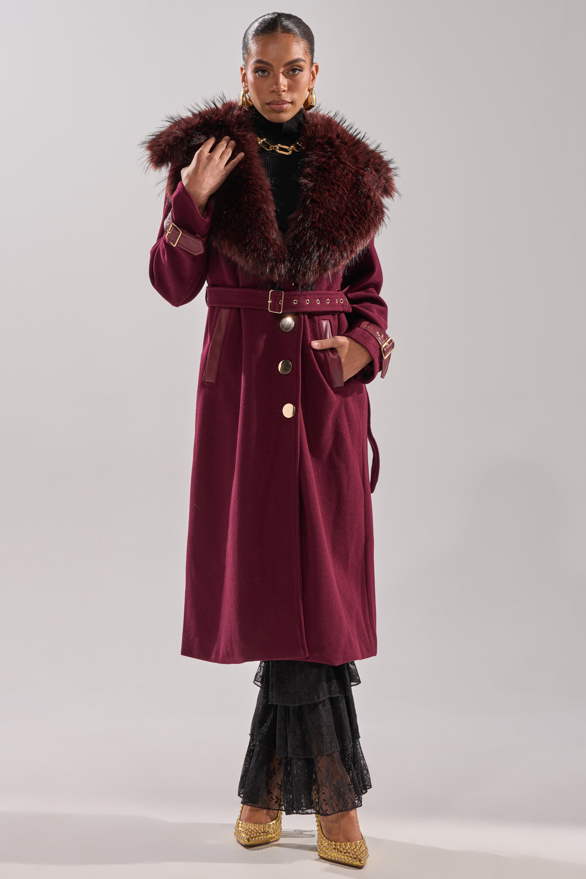 A woman poses against a plain light background wearing the CATALINA FUR COLLAR TRENCH—a longline jacket with a large fur collar, gold buttons, and belt—styled with gold earrings, a black turtleneck, layered black lace skirt, and gold heels. 9