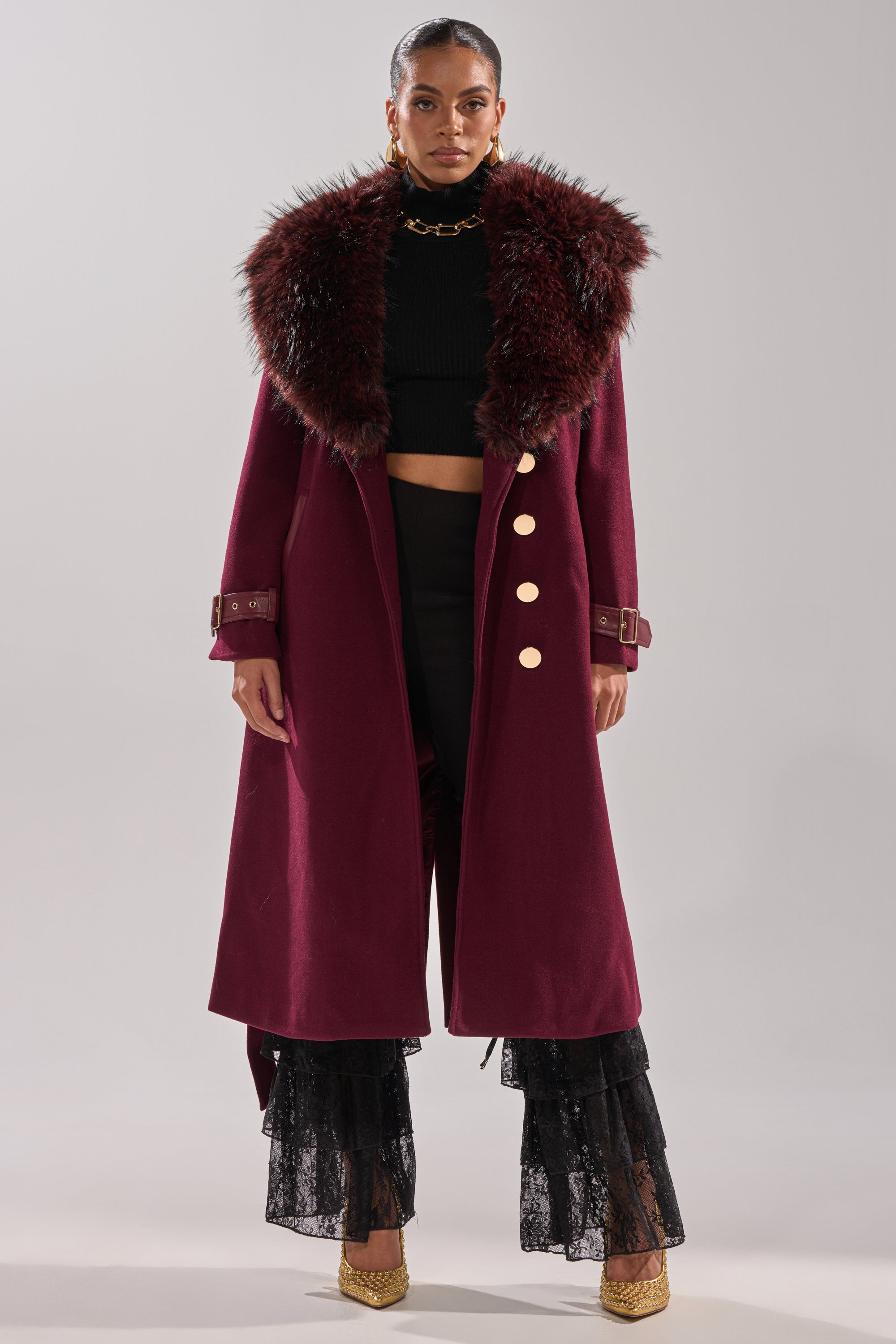 A woman wears the CATALINA FUR COLLAR TRENCH with a maroon faux fur collar and gold buttons, paired with a black cropped top, black lace tiered pants, gold heels, and gold earrings against a plain background. 10