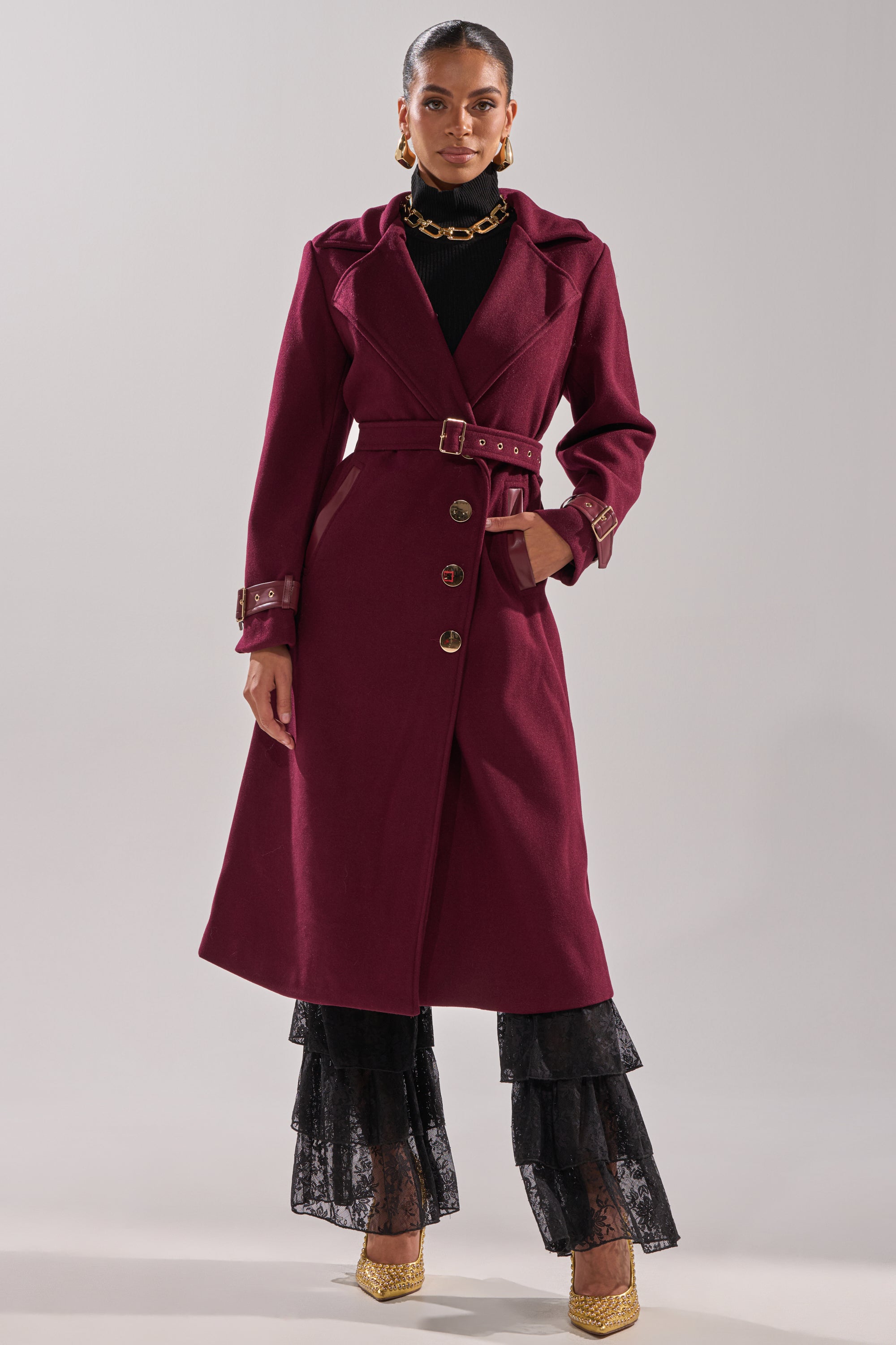 A woman stands against a plain light background wearing the CATALINA FUR COLLAR TRENCH, black lace tiered pants, gold heels, a chunky gold necklace, and gold earrings. 11