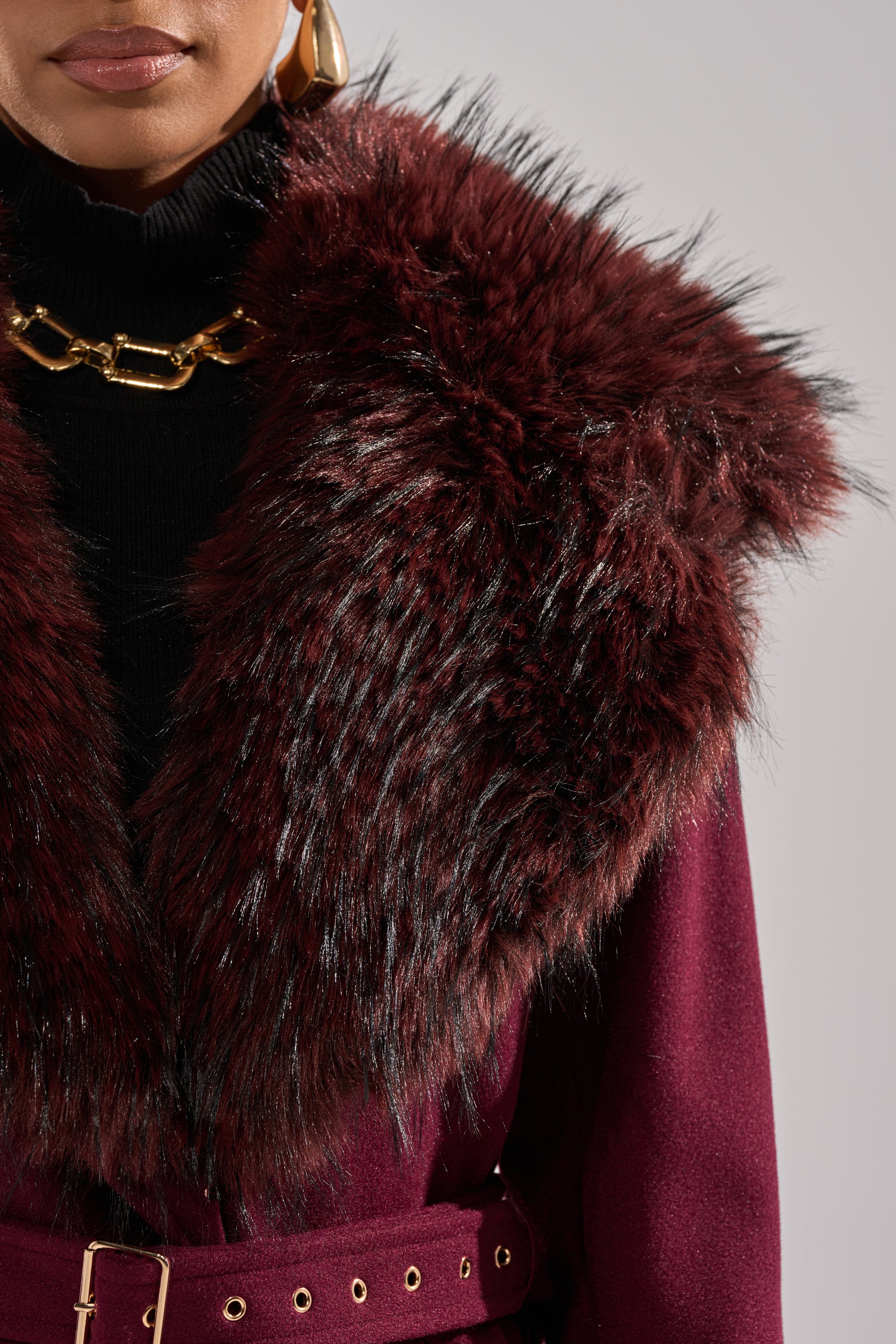 A close-up of a person wearing the CATALINA FUR COLLAR TRENCH in burgundy, styled with a black turtleneck, chunky gold chain necklace, and gold hoop earrings. 12