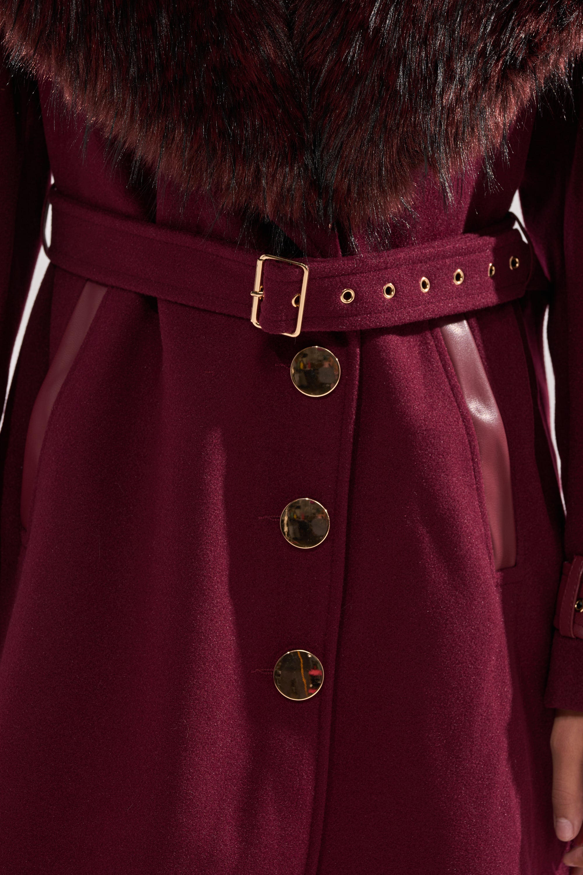 A close-up of the CATALINA FUR COLLAR TRENCH in burgundy, showcasing its wide faux fur collar, gold buttons, matching belt with a gold buckle, and leather-look pocket trim. 13